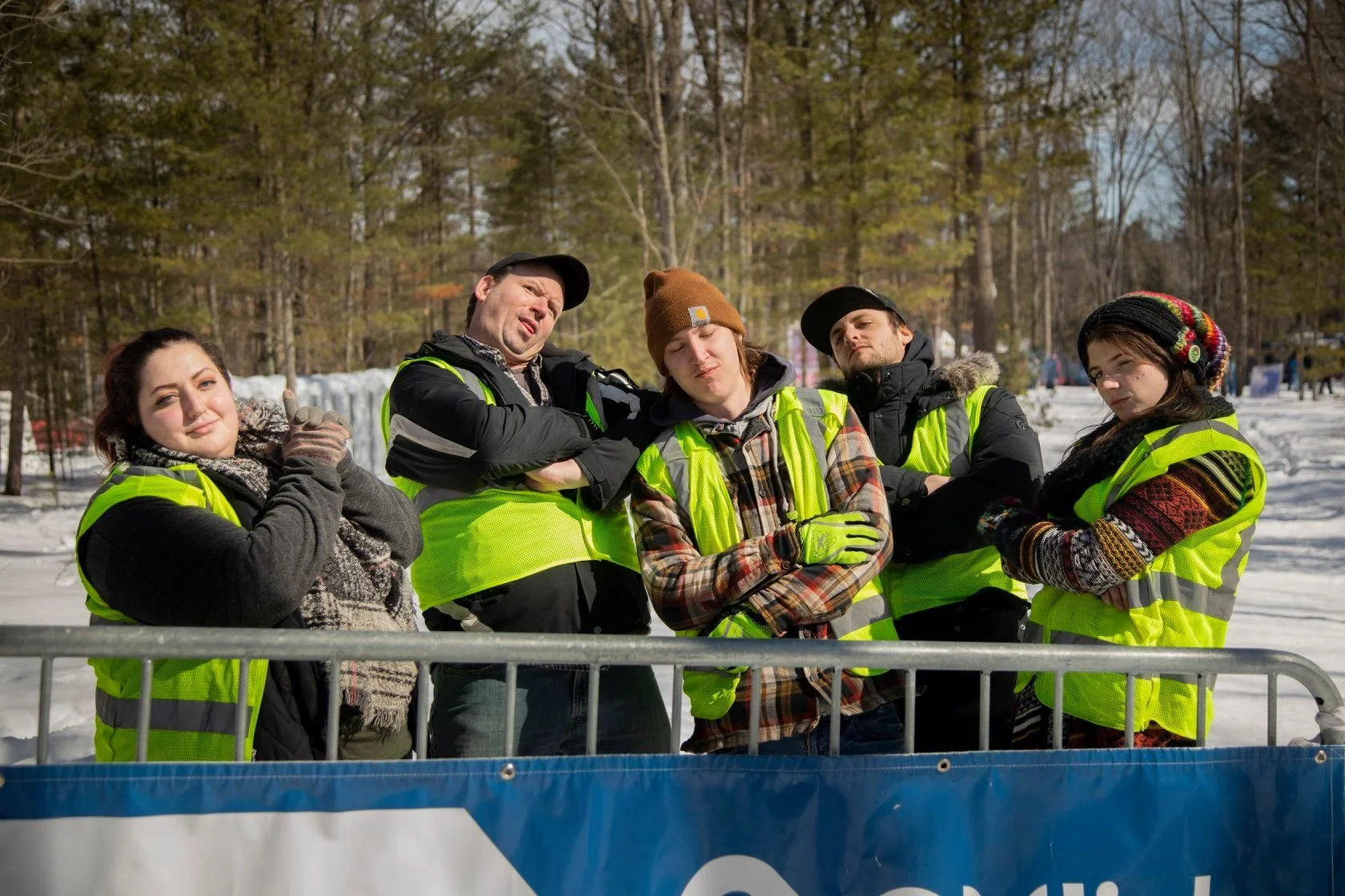 Group of five young people in winter clothing and high-visibility vests posing outdoors on snowy ground, with a forest of trees in the background.