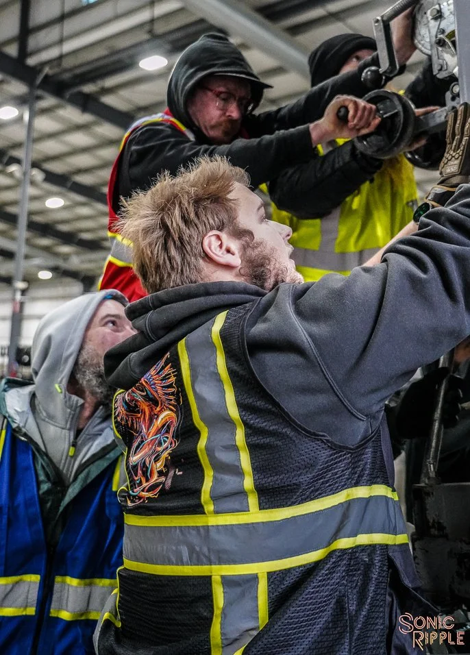 Group of three men working on machinery inside an industrial setting, wearing safety gear and jackets.