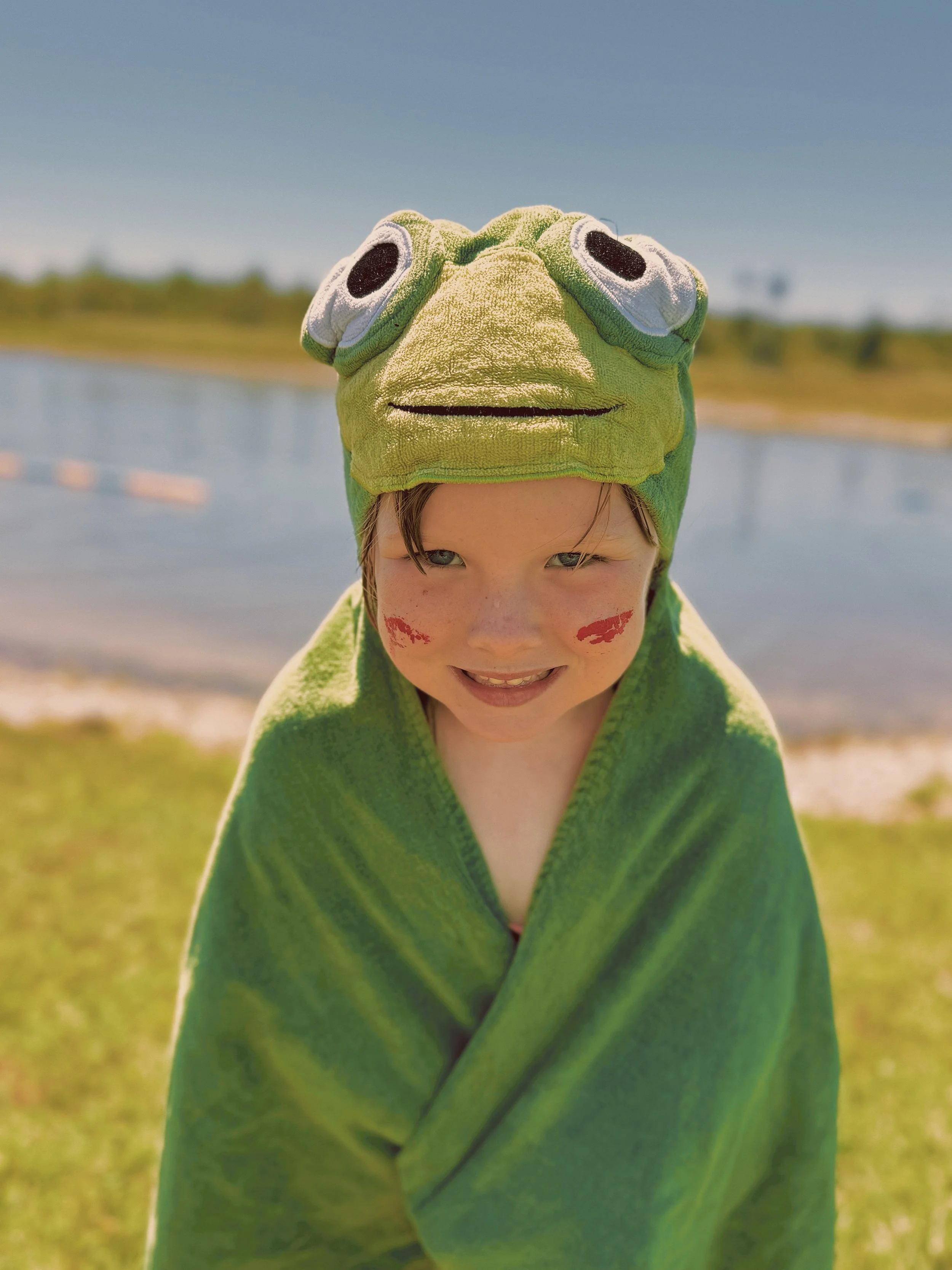 Child dressed in a green towel and wearing a frog towel hat, smiling outdoors by a body of water.