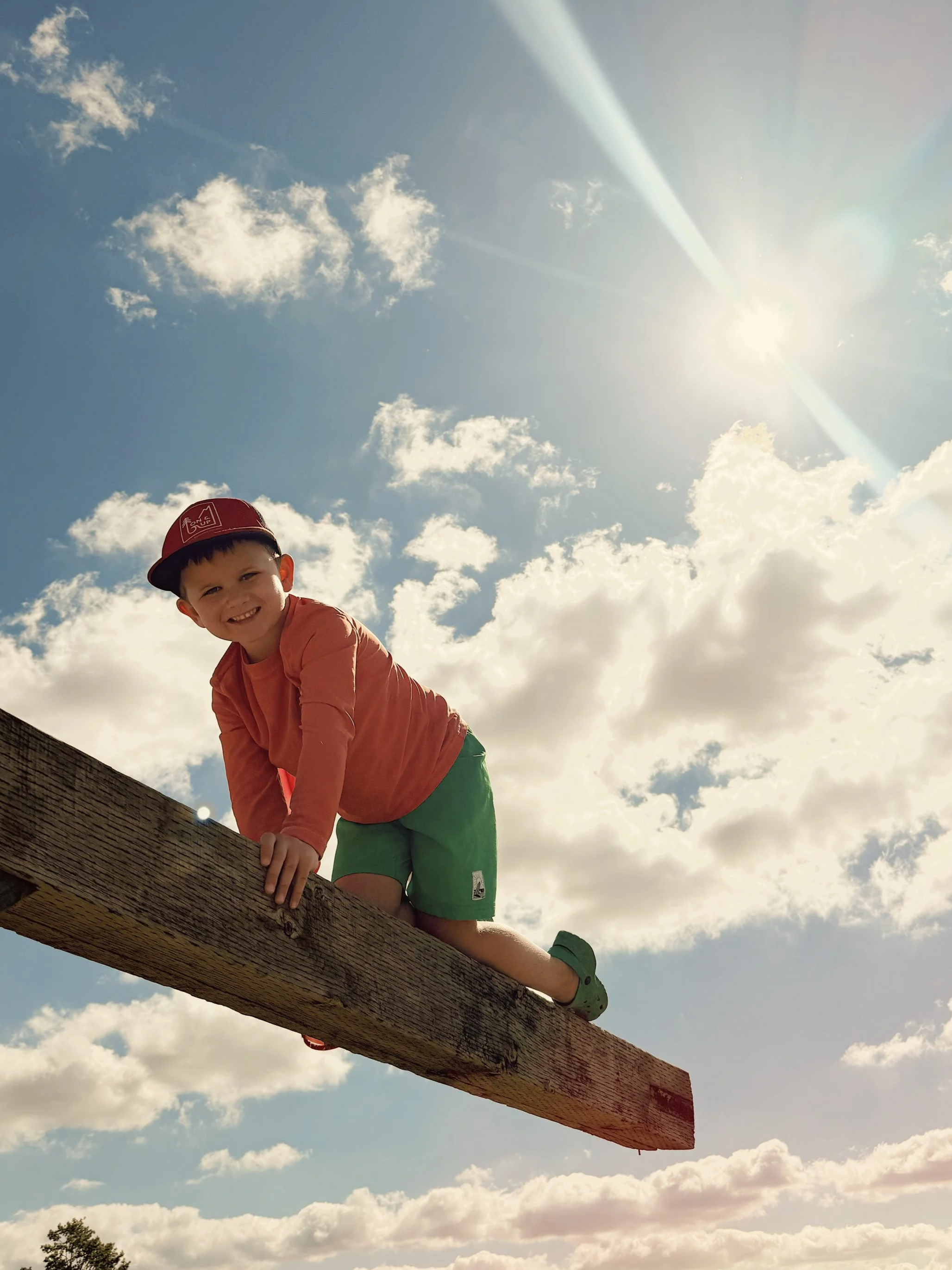 A young boy wearing a red cap, orange long-sleeve shirt, and green shorts balancing on a wooden beam outdoors with a bright sun and partly cloudy sky in the background.