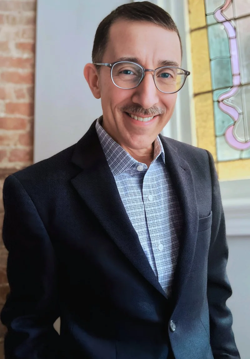 Derek Pollard, founder of Constellar Creative, smiling in a navy blazer and blue checkered shirt, wearing glasses and a neatly trimmed mustache. He is standing in front of a brick wall, with a stained glass window in the background.