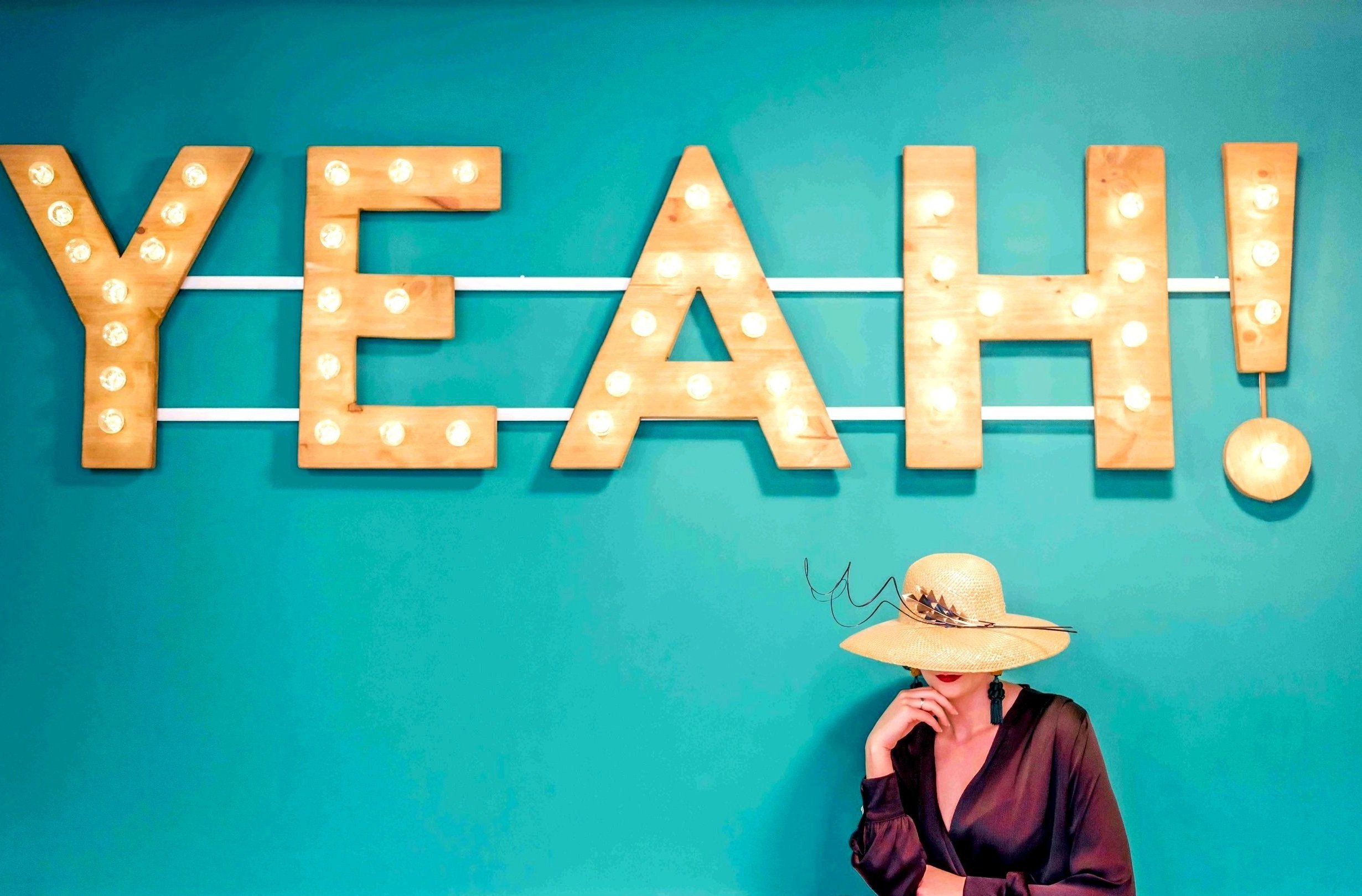 Woman in hat under marquee letters spelling 'YEAH!' on teal wall.