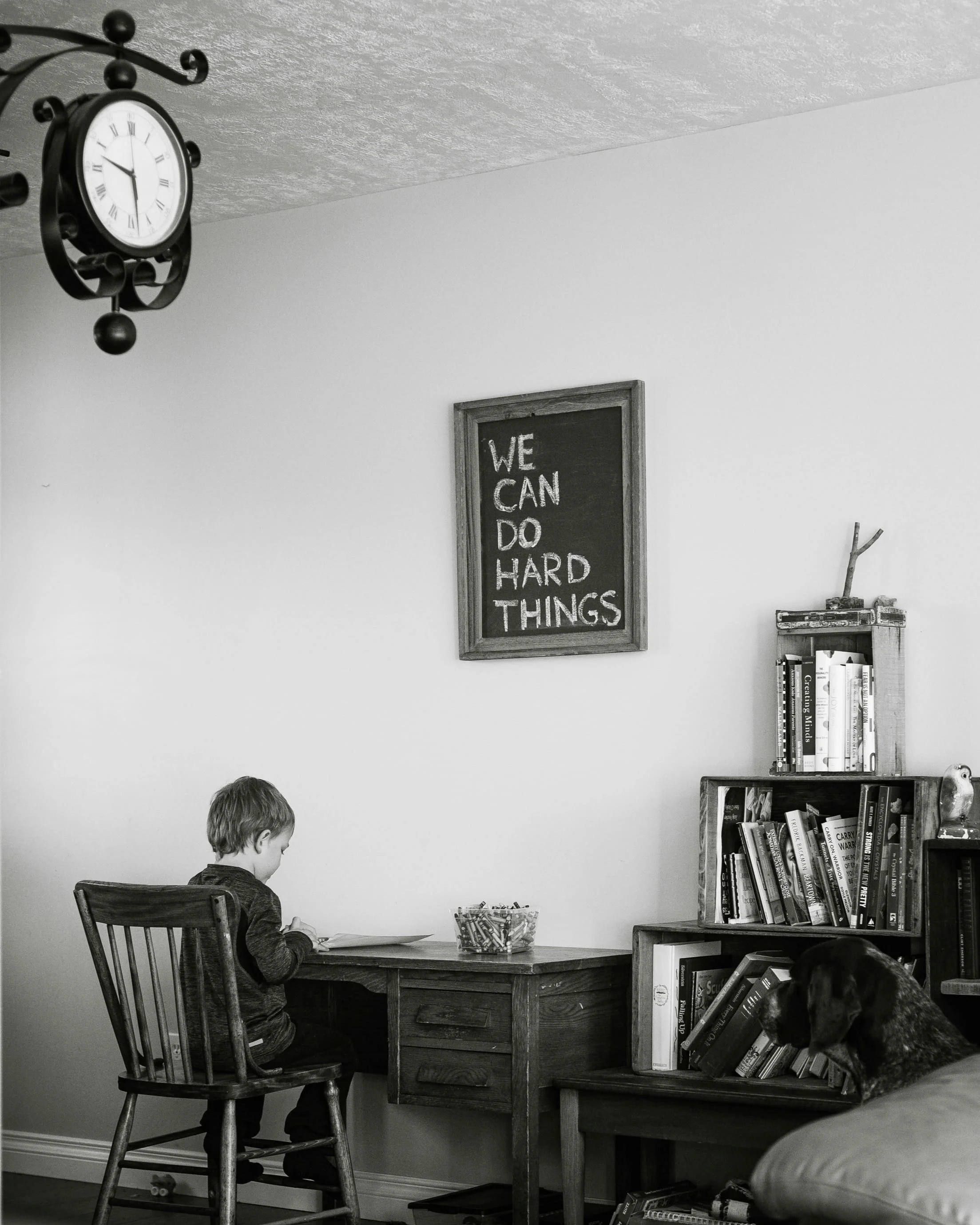 Black and white photo of a young boy sitting at a wooden desk with a basket of colored pencils. A chalkboard on the wall reads "We can do hard things." A bookshelf with various books is nearby, and a dog is in the foreground. A clock is partially visible in the top corner.