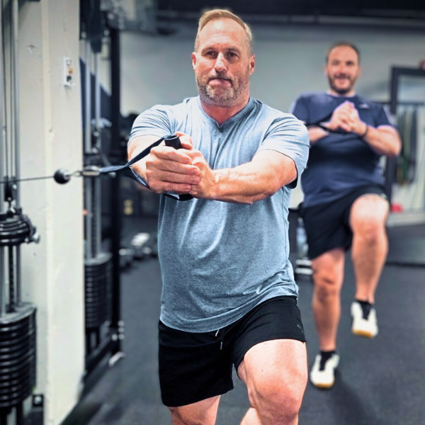 Two men exercising in a gym, one using a resistance band for stretching or strength training and the other in the background with a hand on his hip, both dressed in workout clothes.