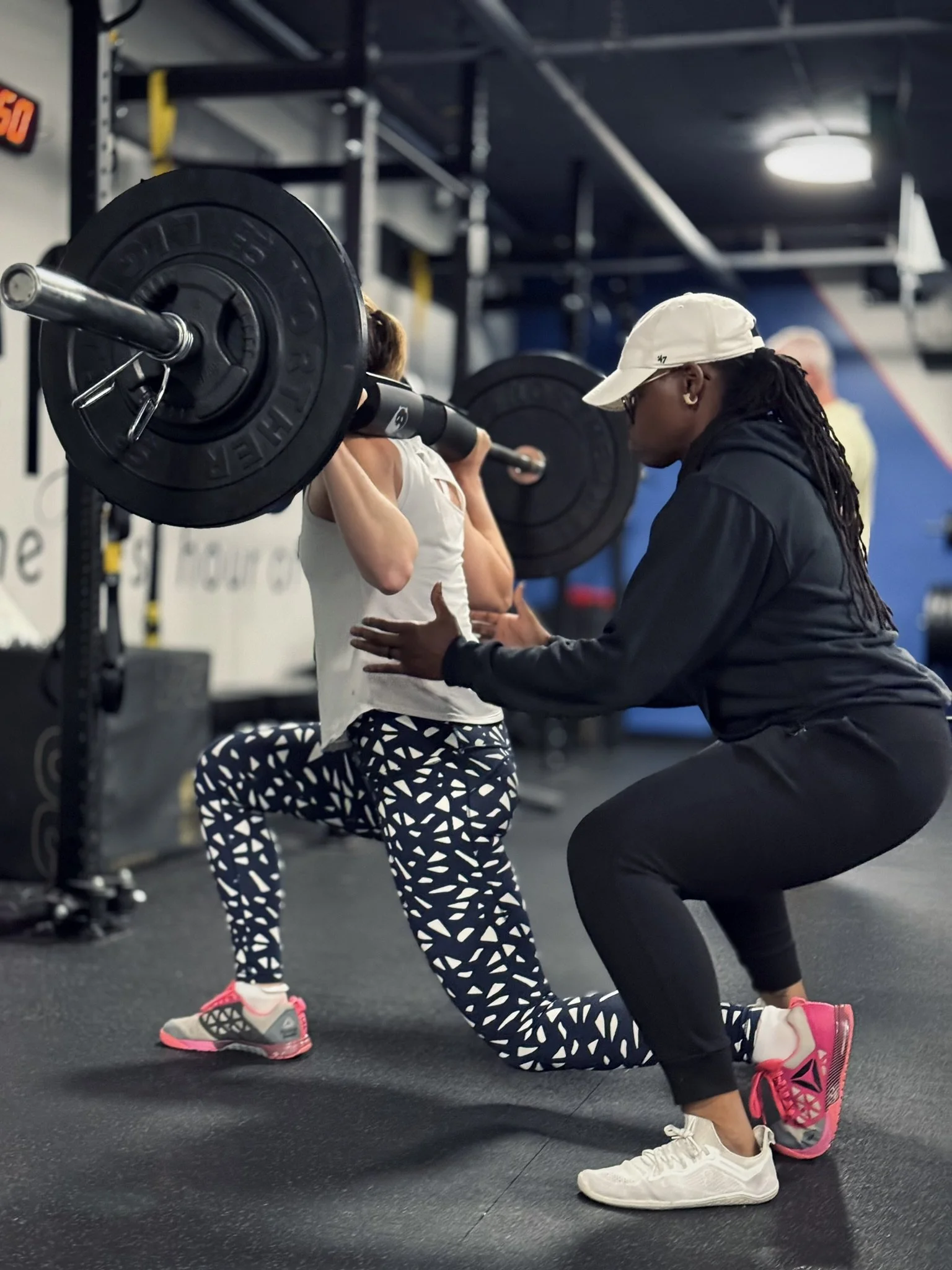 A woman performing a squat with a barbell on her shoulders, supervised by a trainer in a gym.