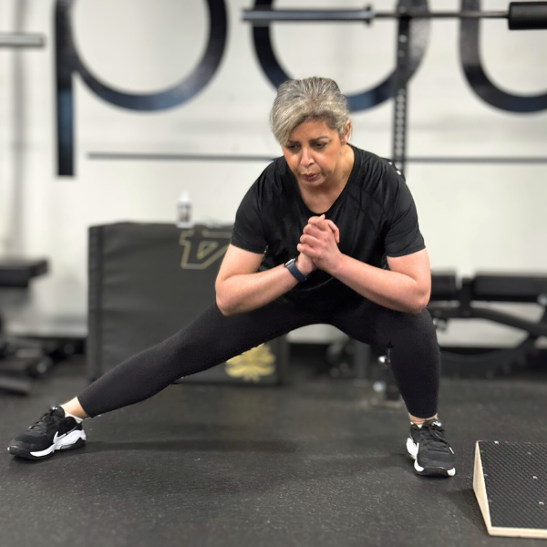 Older woman with gray hair exercising in a gym, performing a wide-legged squat with hands in prayer position.
