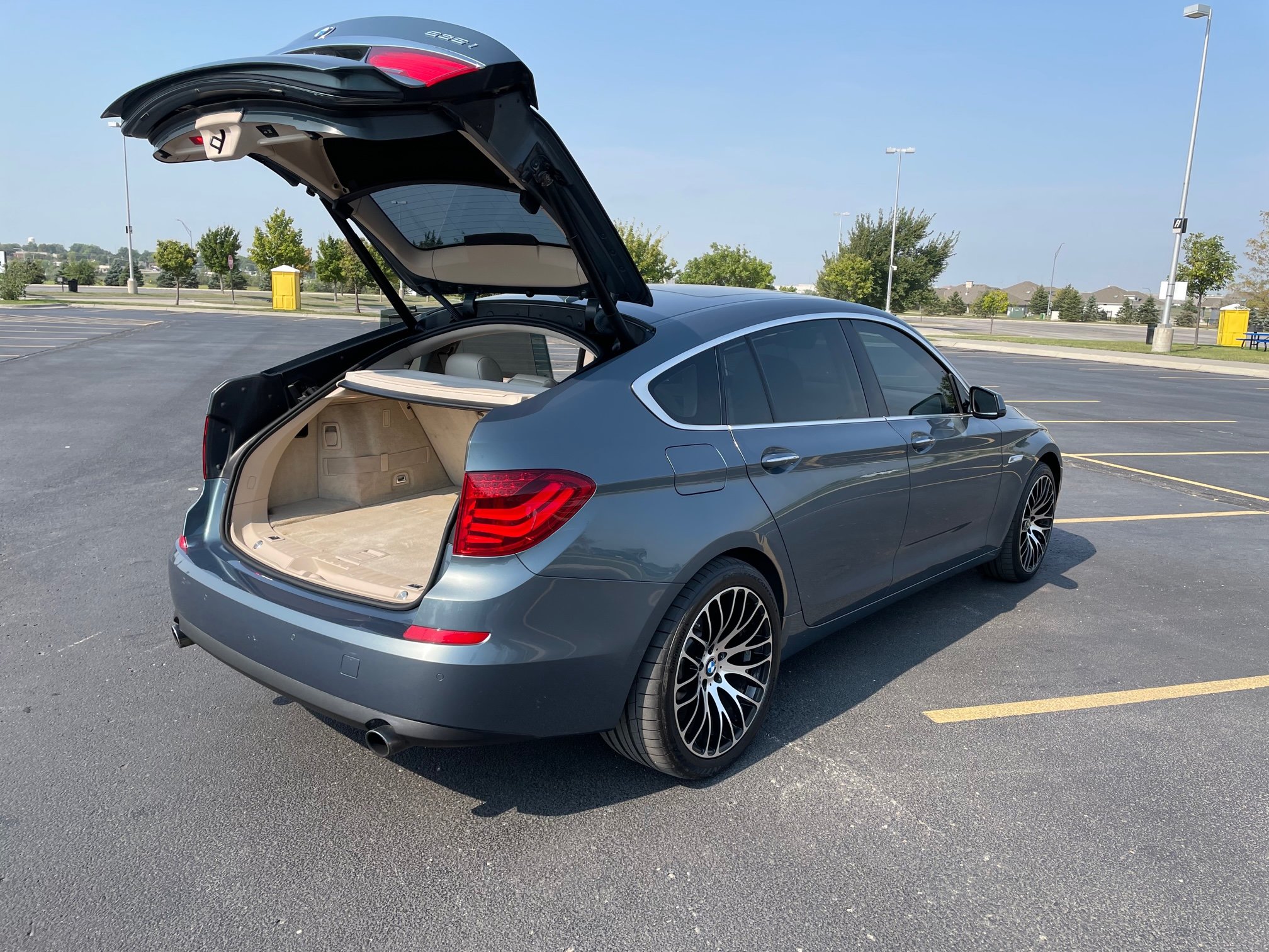 A silver sedan with its trunk open parked in a mostly empty parking lot on a sunny day, with trees and blue sky in the background.
