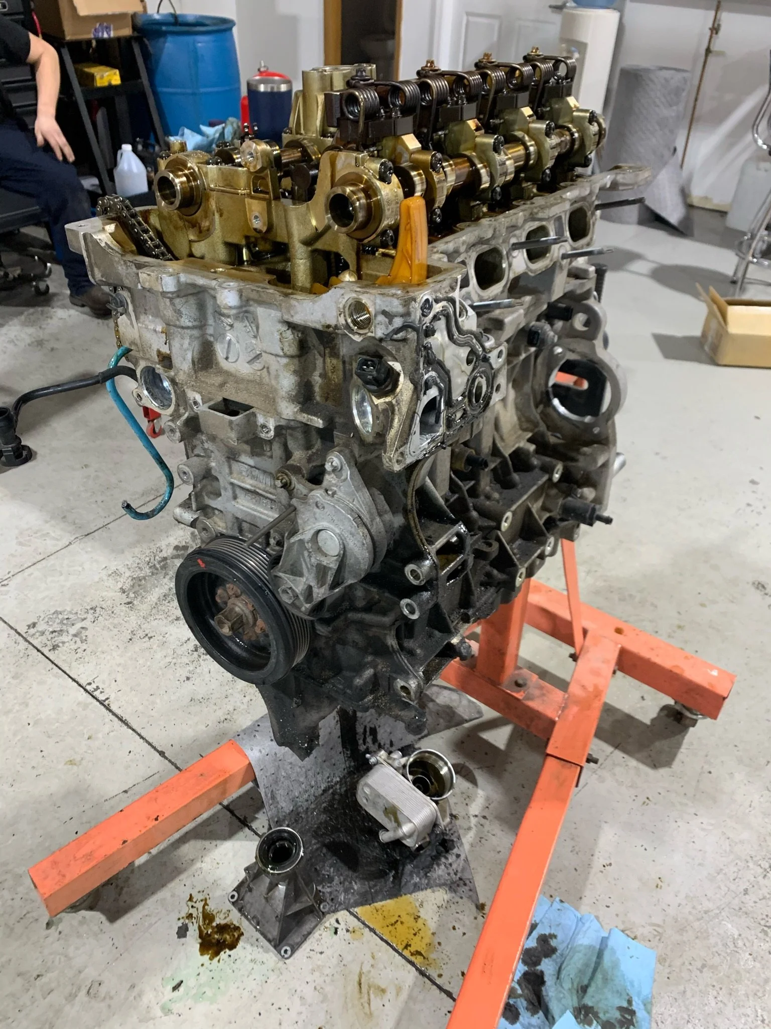 An engine block on a workbench with some engine components, tools, and oil stains in a garage or workshop setting.