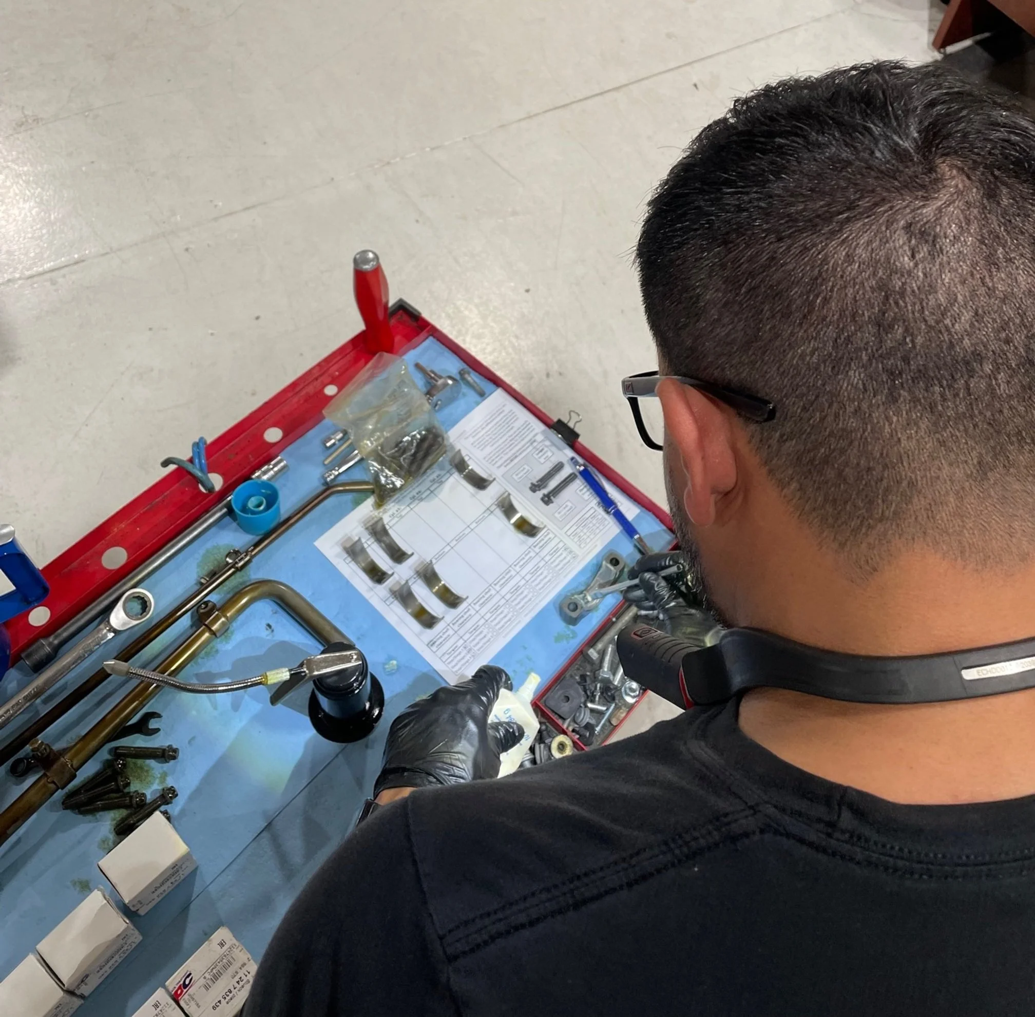 A person with short, dark hair and glasses is sitting on the floor in front of a red toolbox containing various plumbing tools and parts, including pipes, fittings, and boxes of supplies. The person is wearing a black shirt and appears to be working 