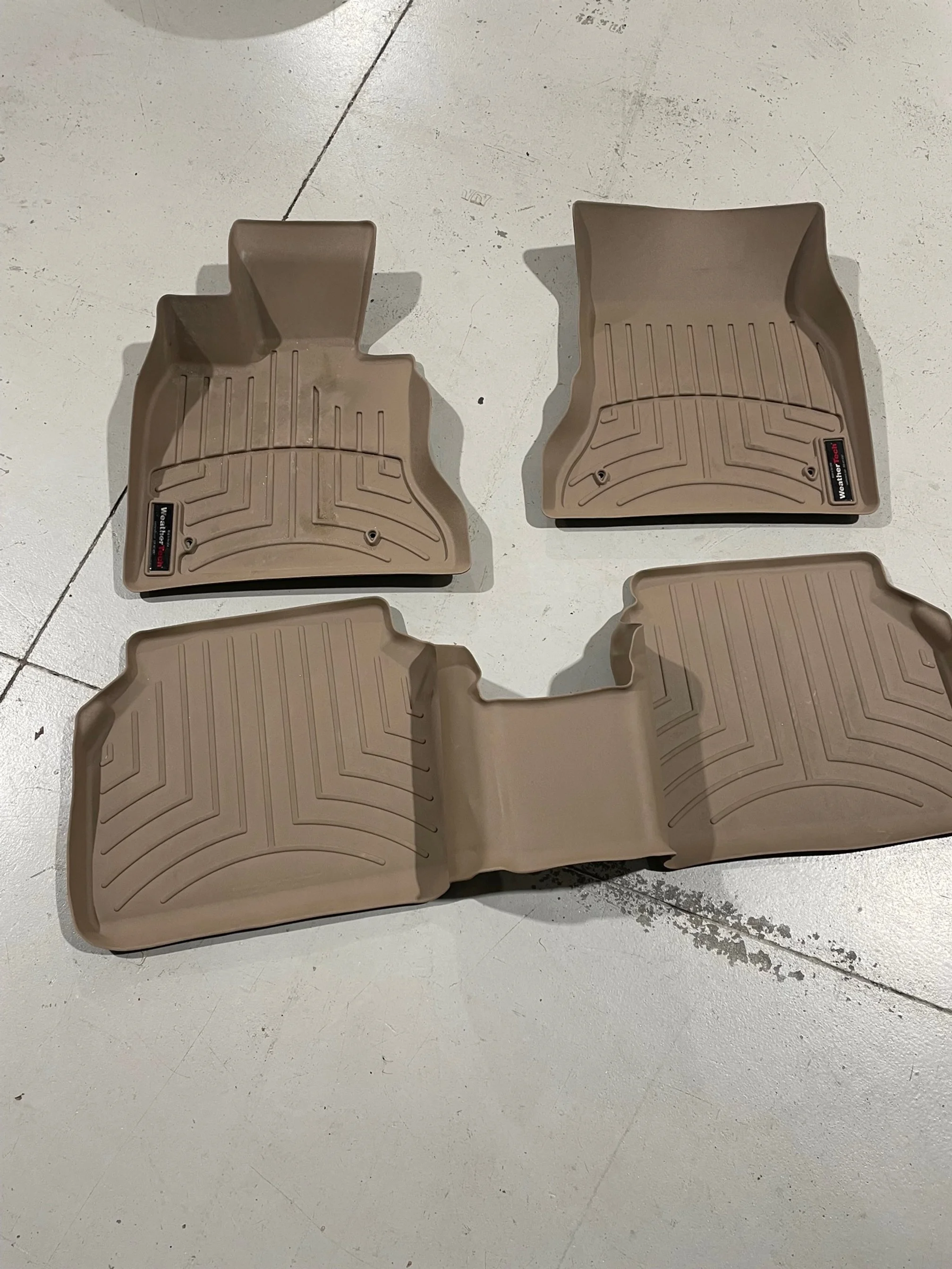 Two beige car floor mats with rubberized grooves and a WeatherTech logo, placed on a white concrete floor.
