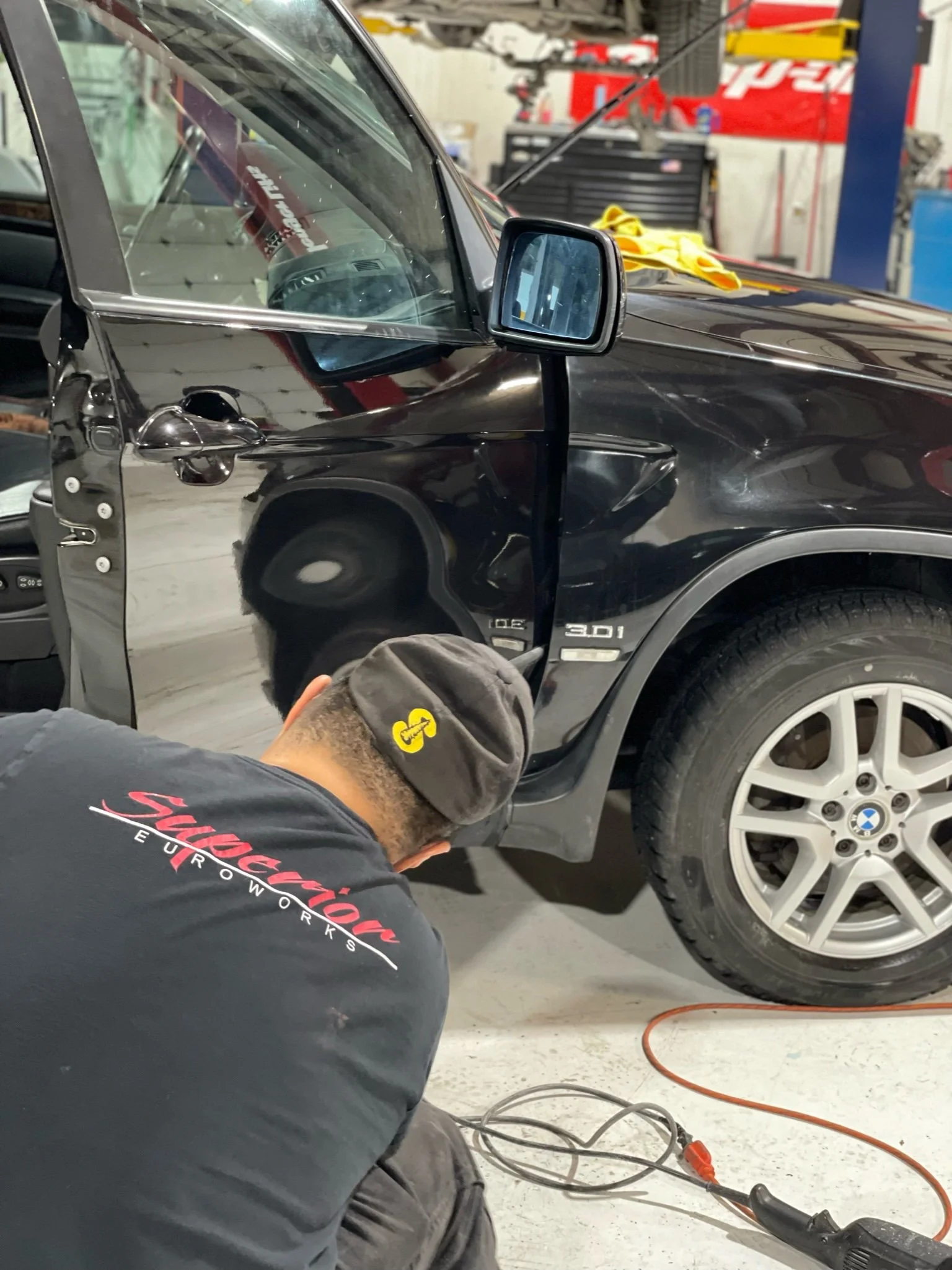 Man working on a black BMW car in a garage or workshop, with tools and equipment visible in the background.