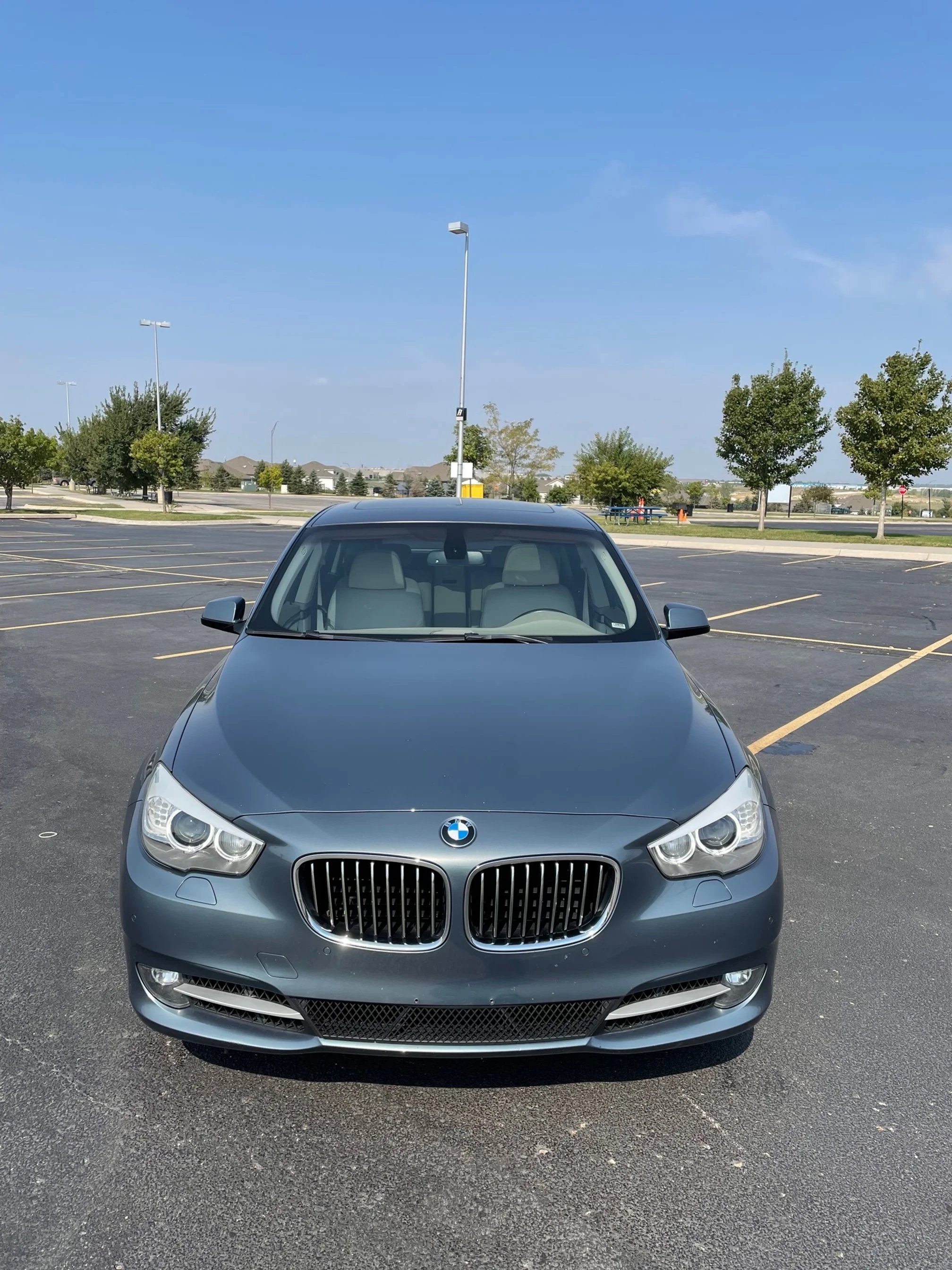Gray BMW car parked in an empty parking lot with trees and a blue sky in the background.