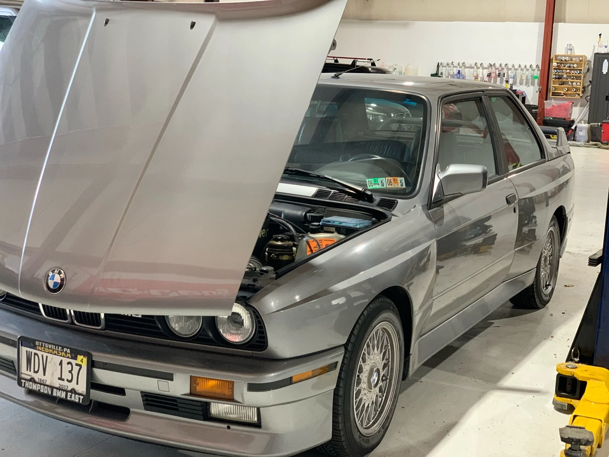 A silver BMW E30 parked inside a garage, with its hood open. The car has Nebraska license plates and BMW logo at the front. Garage shelves with tools and supplies are visible in the background.