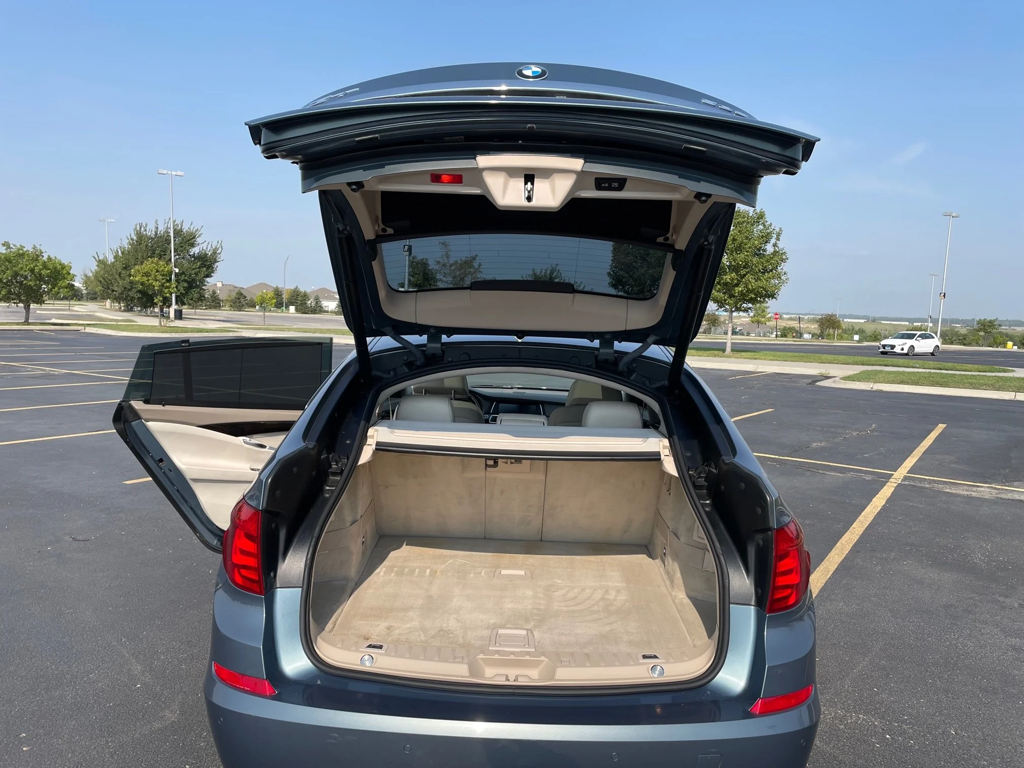 The rear of a silver SUV with an open trunk parked in a mostly empty parking lot under a clear blue sky, with a few trees and a white car in the background.