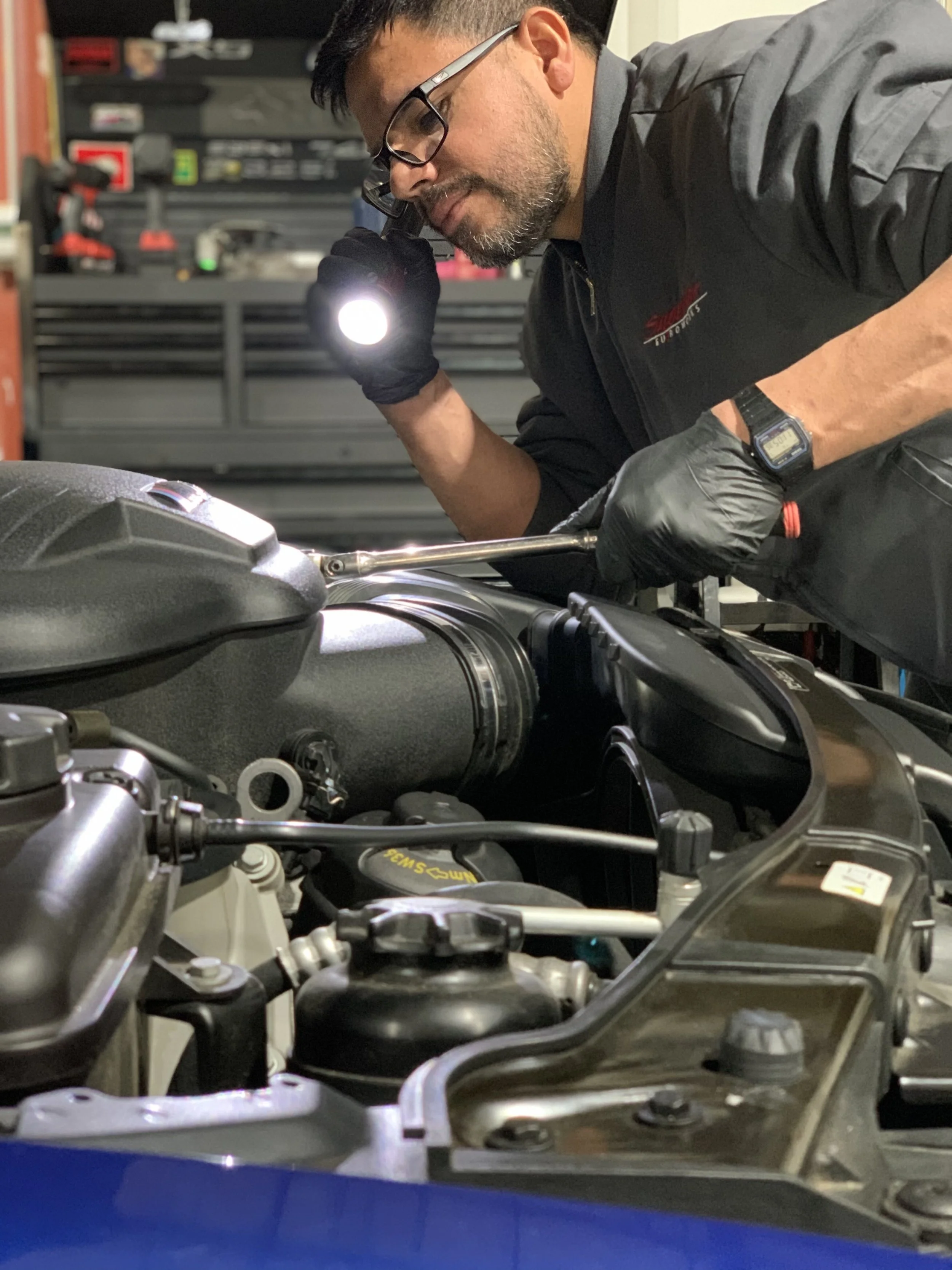 A man wearing glasses and a black jacket with a logo, holding a flashlight, inspecting the engine of a car in a garage.
