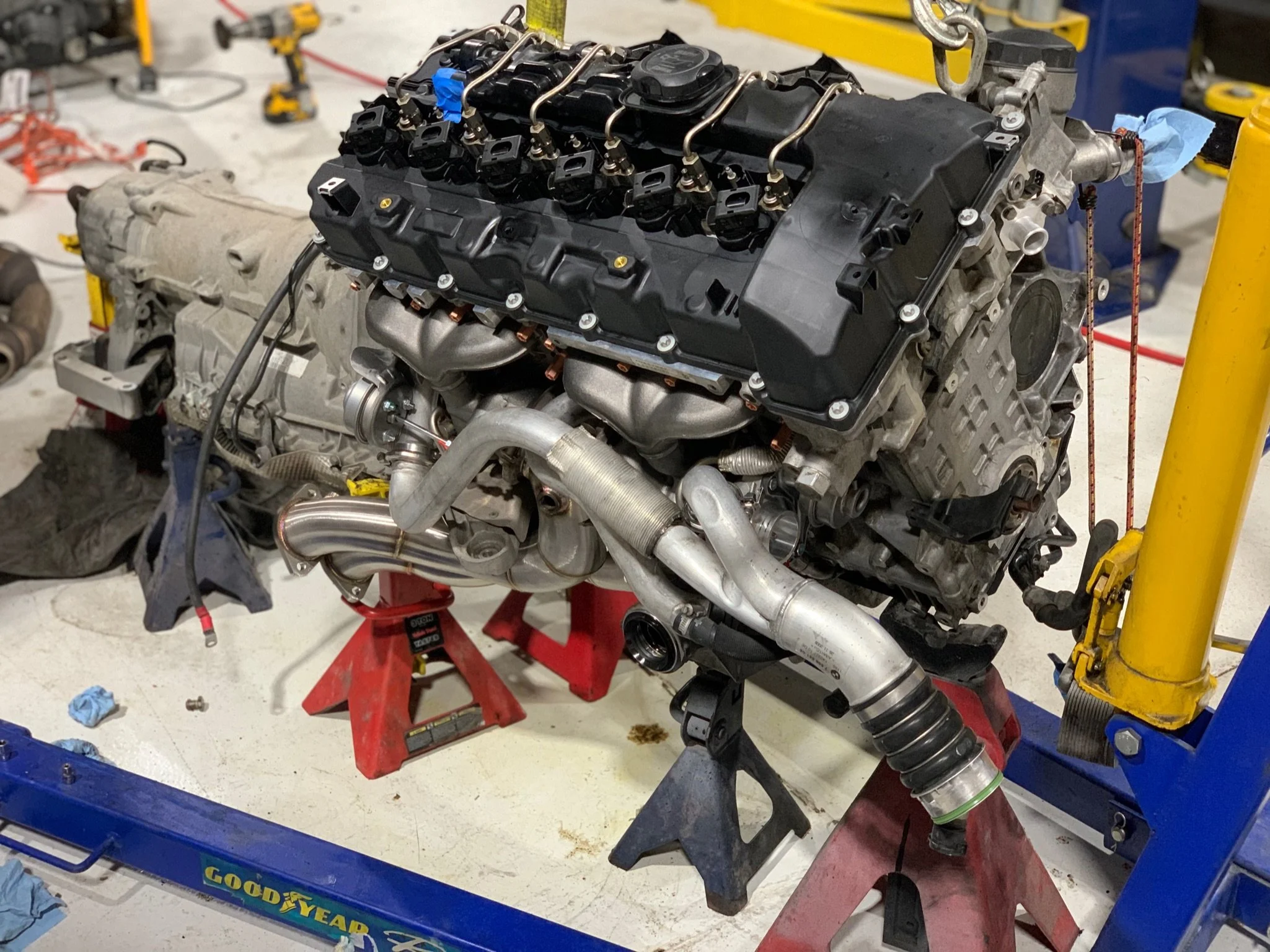 An engine on a workbench in a workshop, with various tools and equipment around. The engine is partially assembled, with a black valve cover and exhaust pipes attached.