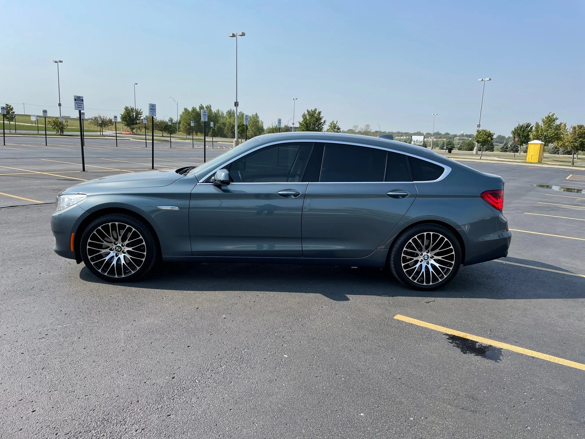 A dark gray BMW sedan parked in an empty parking lot with marked spaces, some trees, and lampposts in the background.