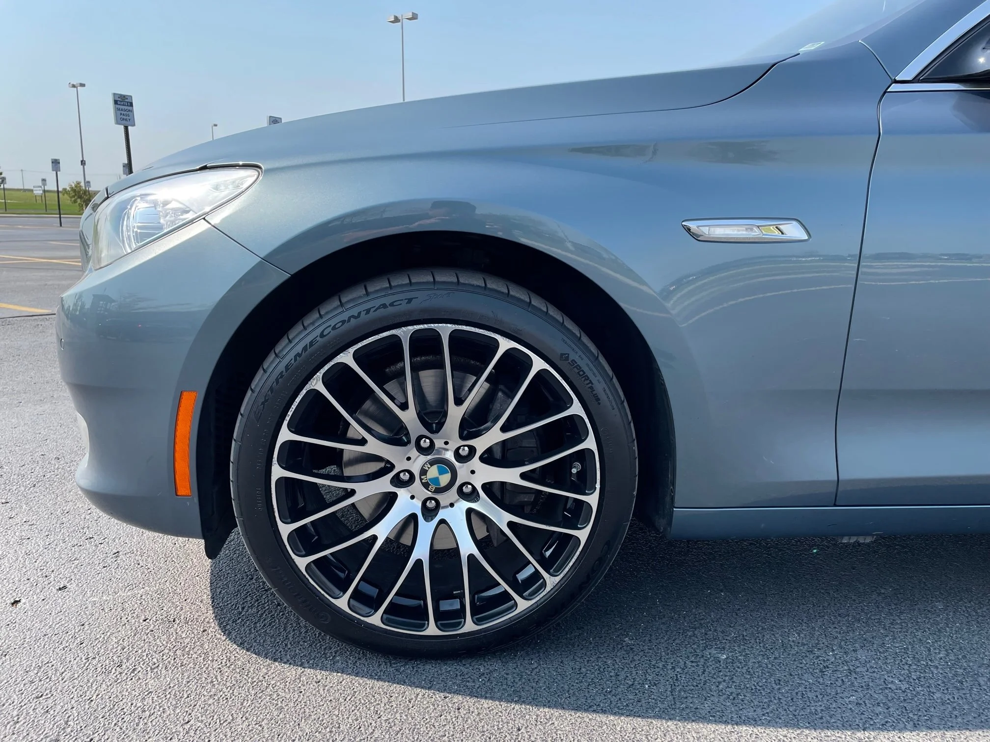 Close-up of the front part of a silver BMW car, showing the headlight, wheel, and part of the hood in a parking lot.