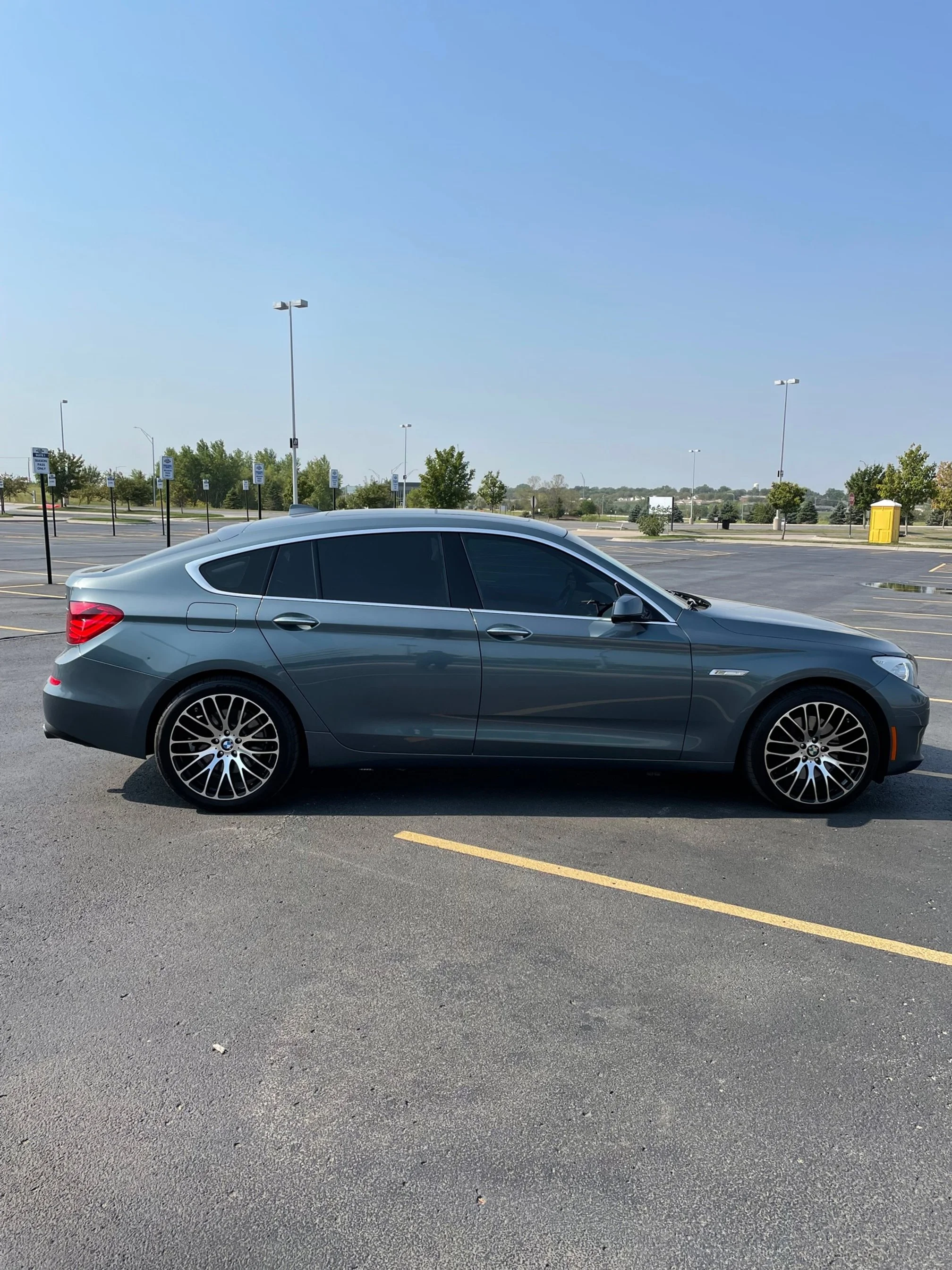 A silver sedan parked in an empty parking lot under a clear blue sky with some trees and lampposts in the background.