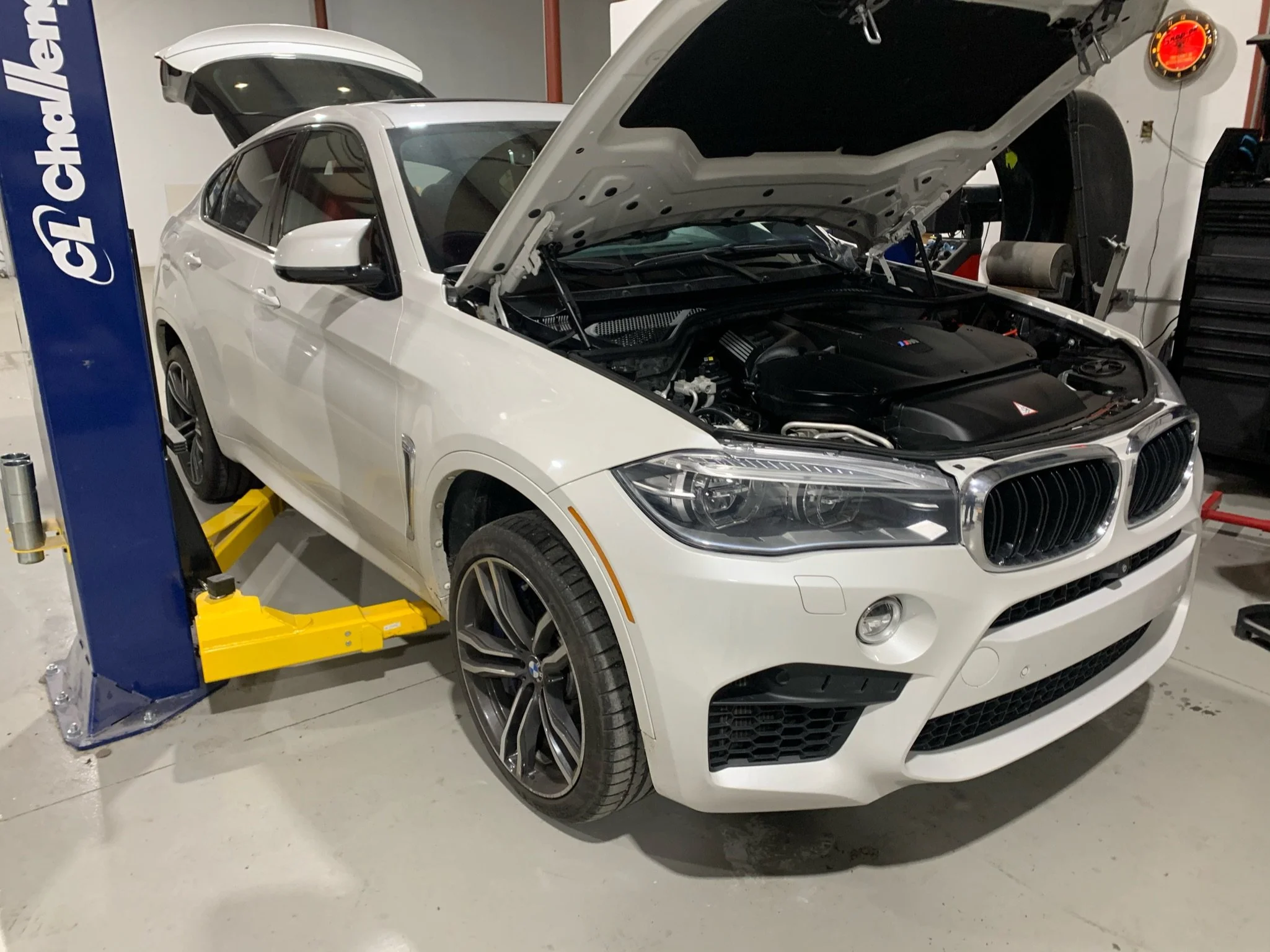 A white BMW car with its hood open, in a car repair or service garage. The garage has an automatic lift, tools, and equipment visible in the background.