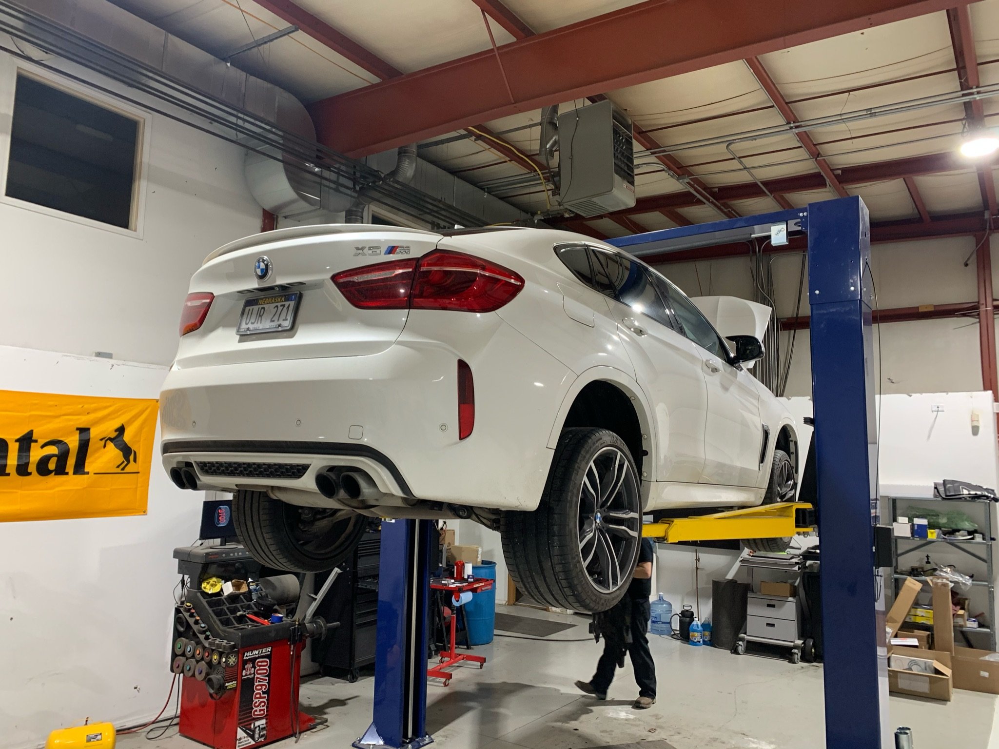 A white BMW X5 M SUV is elevated on a blue hydraulic lift inside a garage or automotive workshop. The garage has a high ceiling with exposed red metal beams and ventilation ducts. There are tools and equipment around, including a tool cart, a yellow 