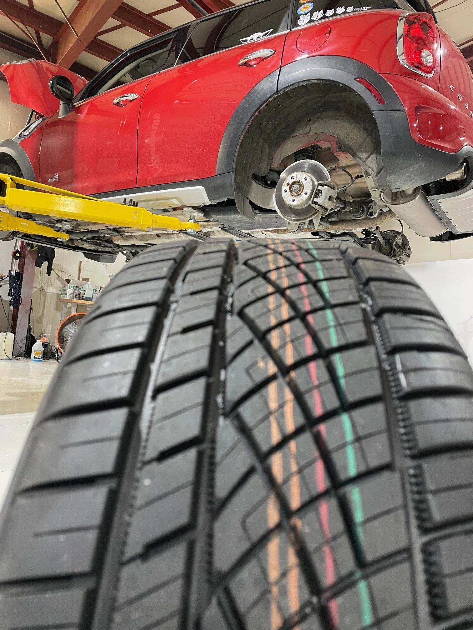 A red Mini Cooper car is lifted on a hydraulic lift in a garage, with its front wheel removed, exposing the brake assembly and suspension components.