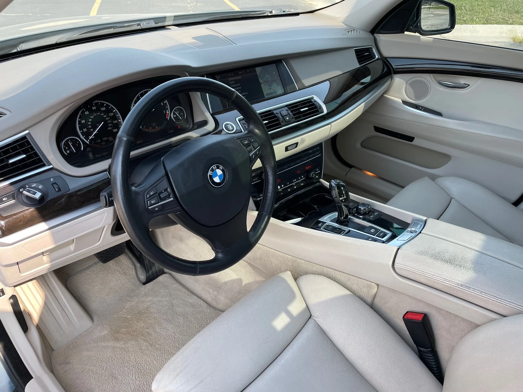 Interior of a BMW vehicle showing the steering wheel, dashboard, and front seats. The dashboard features a navigation screen and control buttons, with beige and black interior accents.