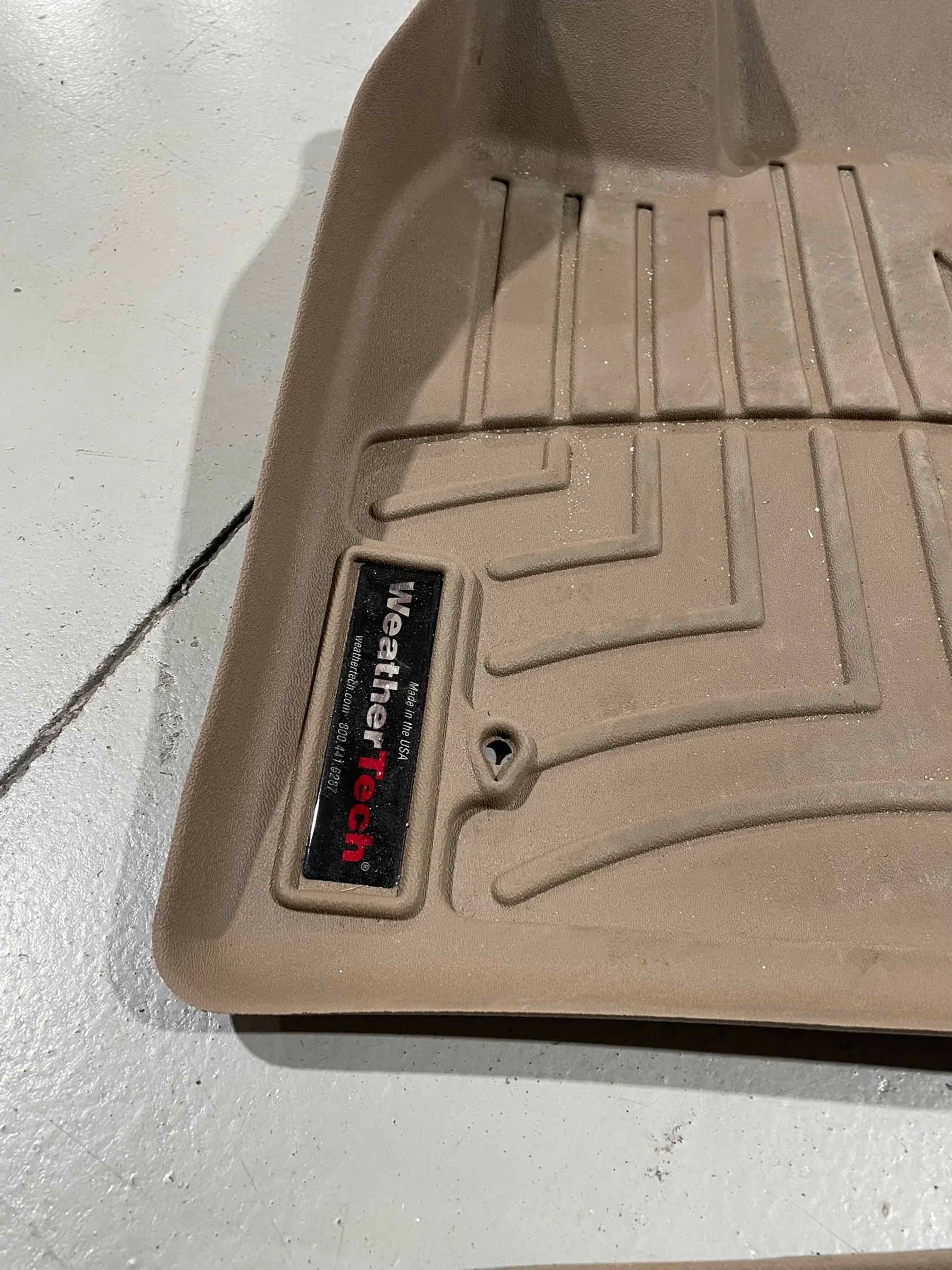 Close-up of a beige WeatherTech floor mat with grooves and a label, placed on a concrete surface.