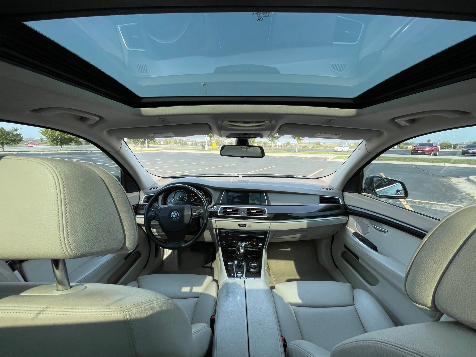 The interior of a luxury BMW vehicle showing beige leather seats, the steering wheel, dashboard, and panoramic sunroof with a view of a parking lot outside.