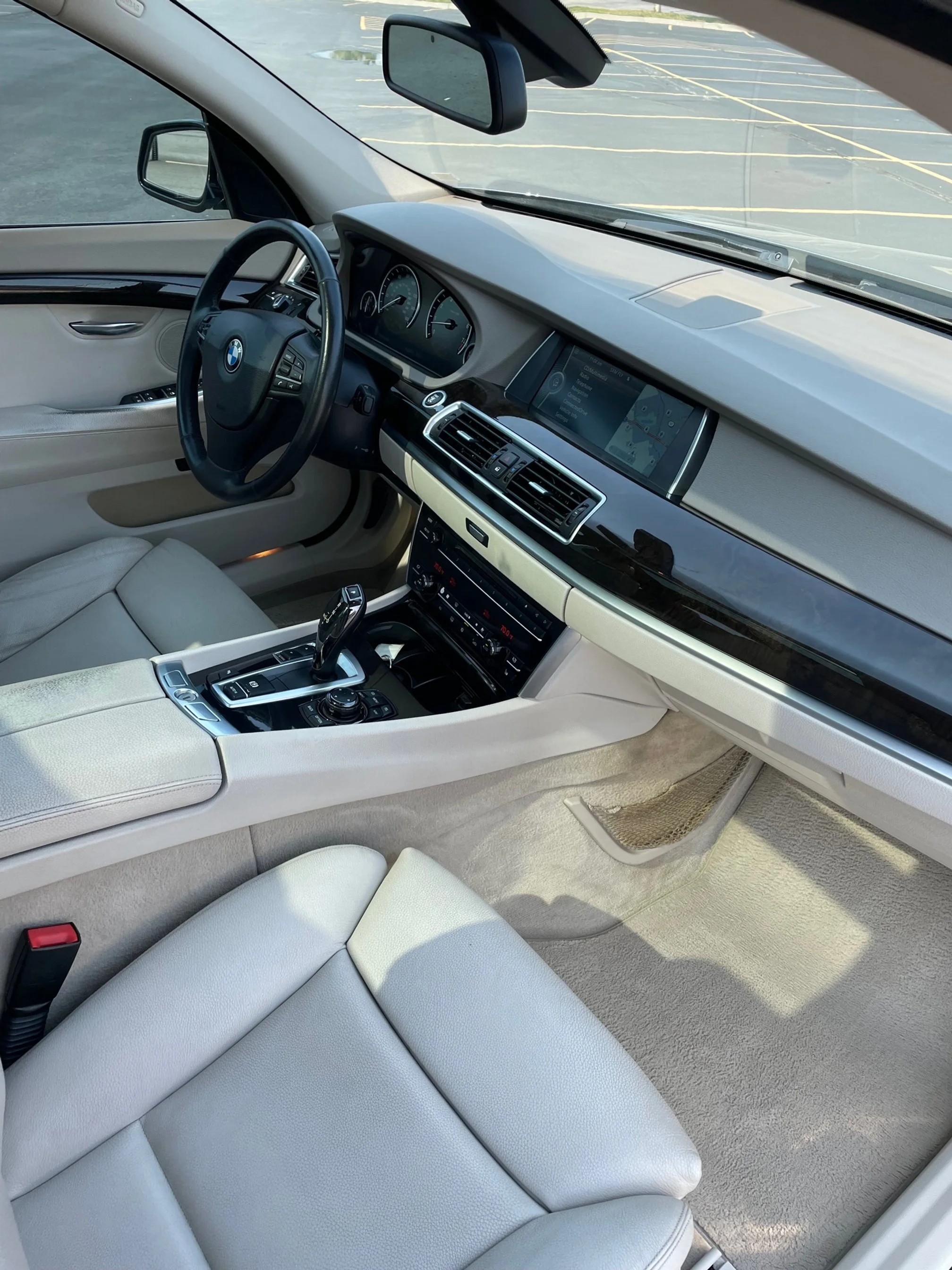 Interior of a BMW vehicle showing a light-colored leather seat, dashboard with speedometer, and steering wheel with BMW logo, parked in a lot with yellow parking lines visible through the windshield.