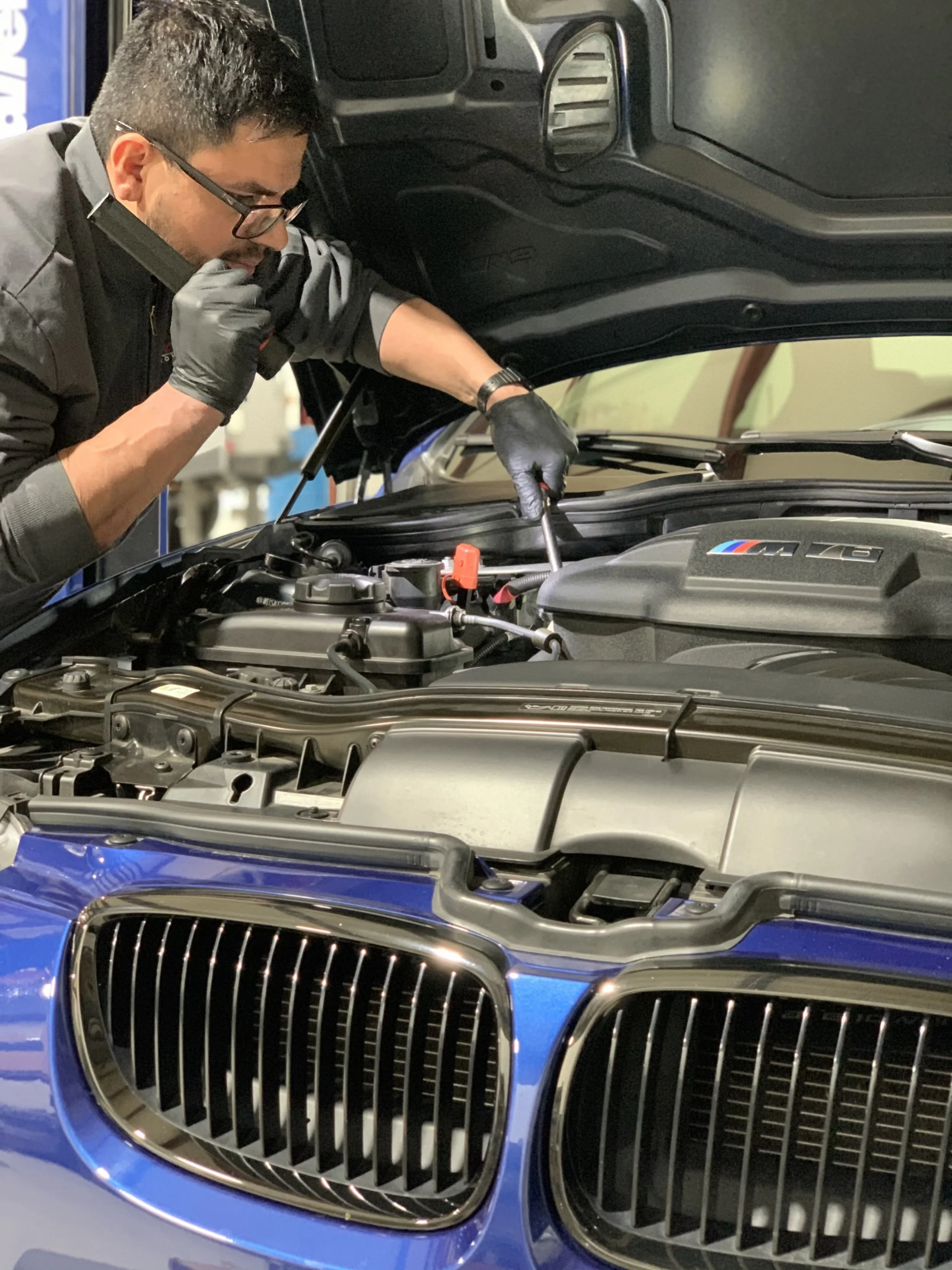 A man wearing glasses, a black jacket, and black gloves is inspecting or working on the engine of a blue car with an open hood inside a garage or workshop.