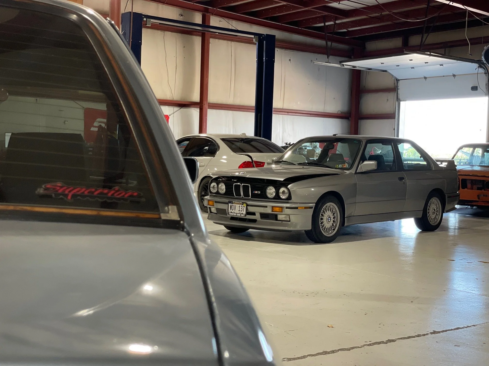 View of a garage interior with three cars parked inside, including a silver BMW E30 in the foreground, a white car behind it, and a partially visible darker car on the left. The garage has a high ceiling with exposed beams and a partially open garage