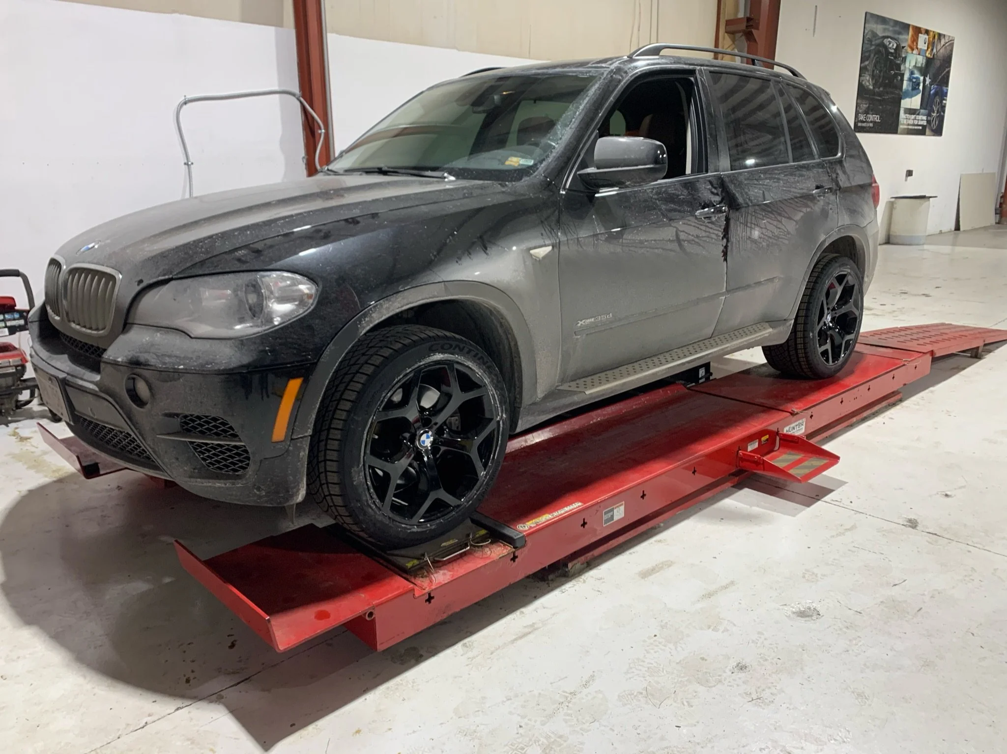 A black BMW SUV parked on a red automotive lift inside a garage or workshop, with posters on the wall behind it.
