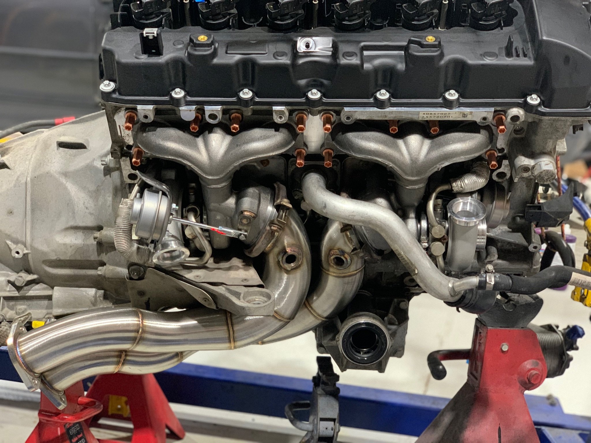 Close-up of a car engine mounted on red stands, showcasing the engine block, intake manifold, and exhaust header pipes.