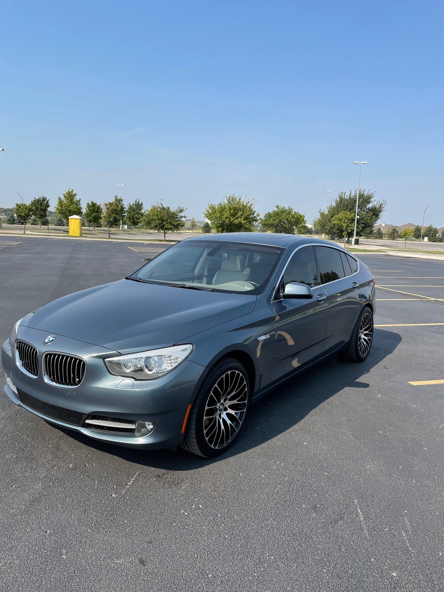 A silver BMW car parked in a mostly empty parking lot on a sunny day, with a row of trees and a yellow utility box in the background.
