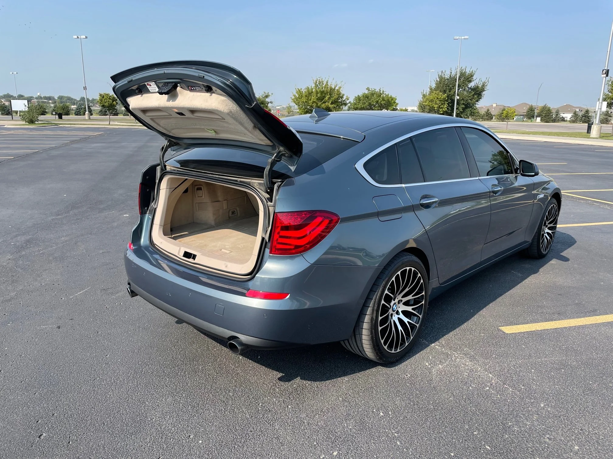 A gray luxury sedan car with an open trunk in an empty parking lot under a clear blue sky.