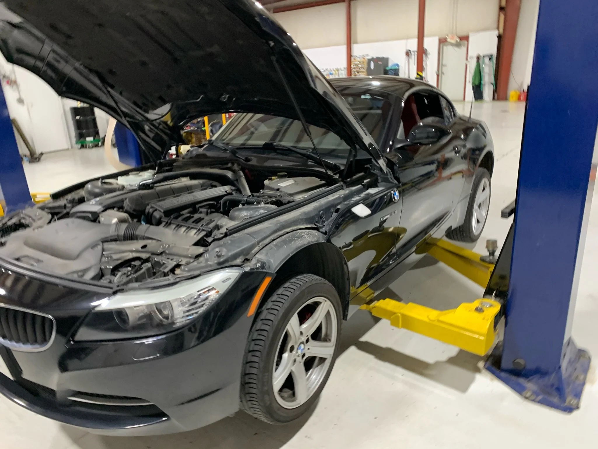 Black BMW sports car with the hood open, parked inside a garage or workshop with various tools and equipment in the background.