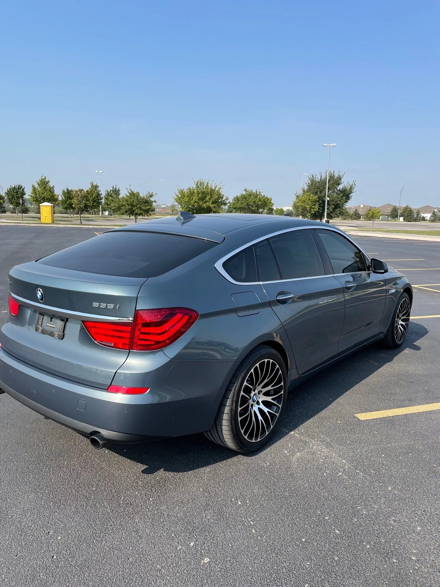 A silver BMW sedan parked in an empty parking lot with trees, streetlights, and a yellow utility box under a clear blue sky.