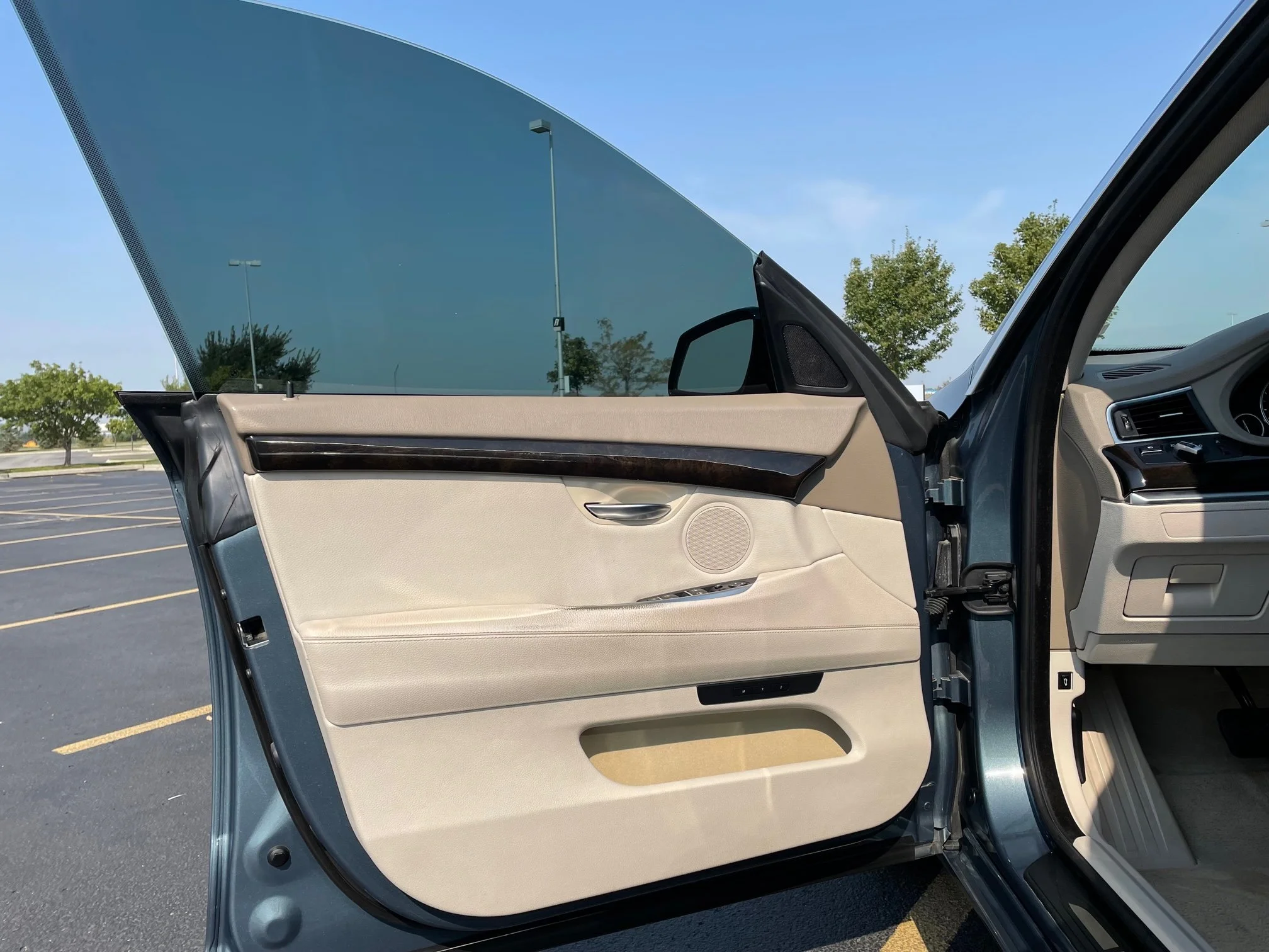 Inside a car, passenger side door open, showing beige interior, control panel, air vent, and windows with view of parking lot and trees outside.