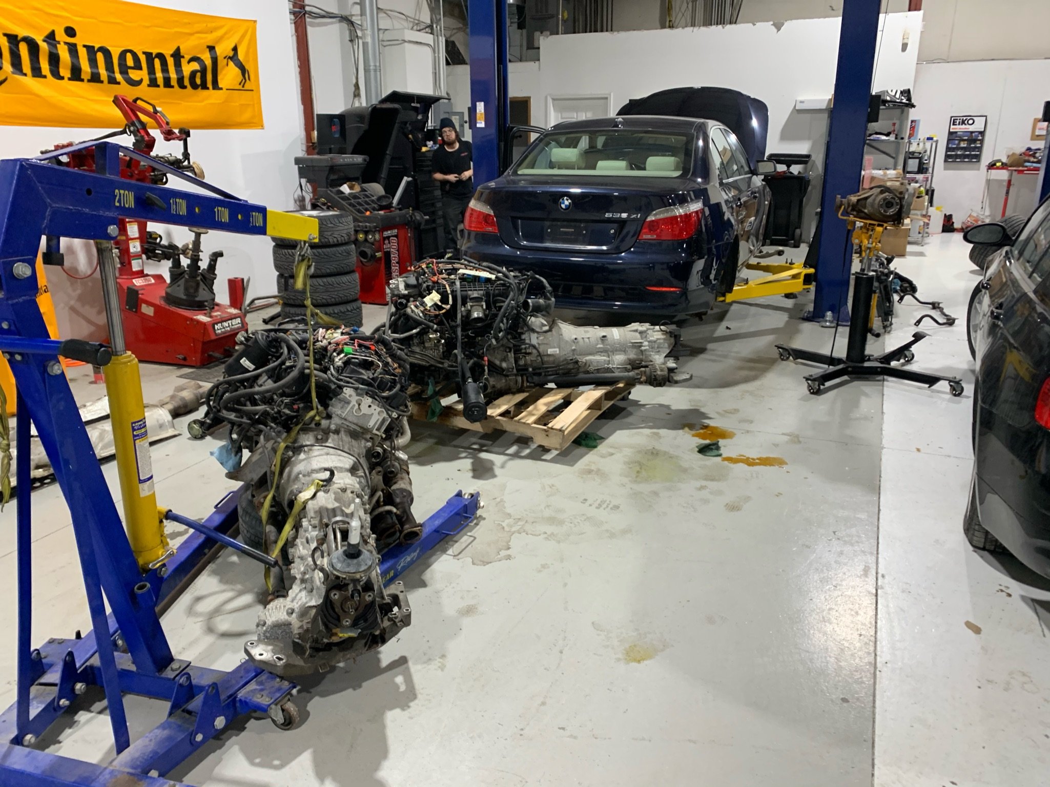 An automotive repair shop with a black BMW parked on a lift, disconnected engine, and various tools and tires around. A mechanic is seen working in the background near a computer, and an engine hoist is in the foreground.