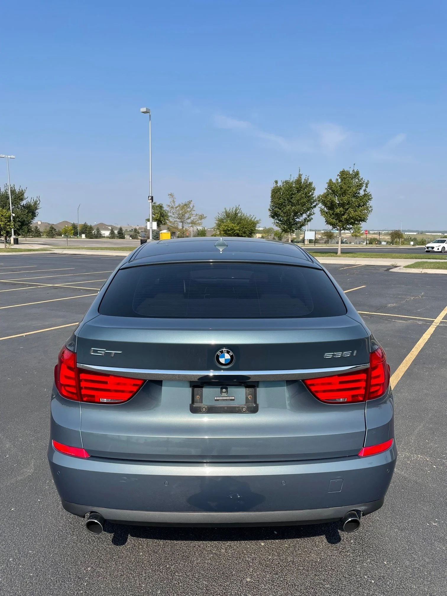 Rear view of a silver BMW 3 Series sedan parked in an empty parking lot with trees, light poles, and a blue sky in the background.
