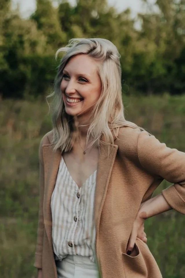 A smiling woman with blonde hair standing outdoors in a field with trees in the background, wearing a beige coat over a striped button-up shirt.