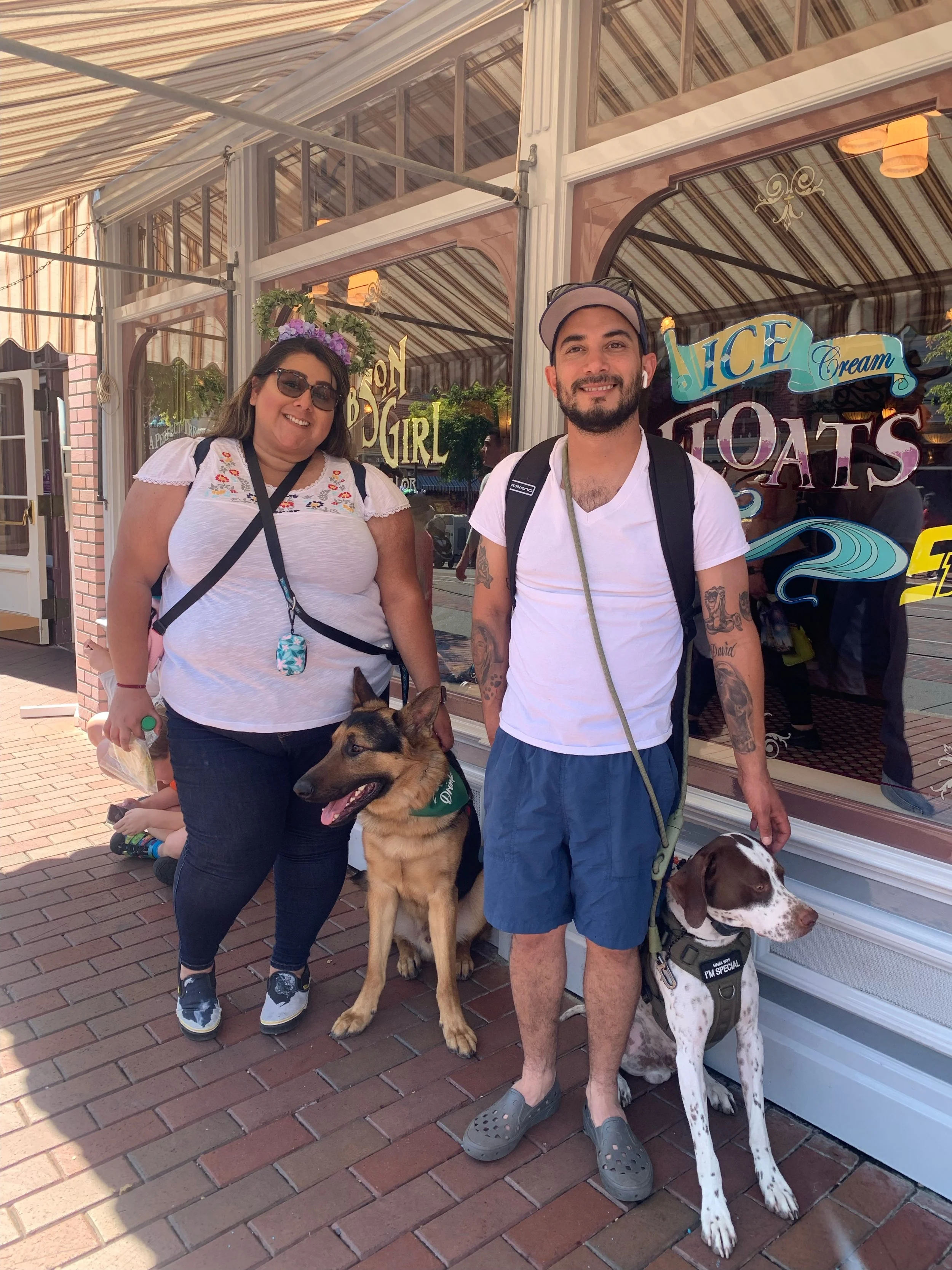 A woman and a man with two dogs standing outside an ice cream shop. The woman is wearing sunglasses, a white embroidered top, and dark jeans, holding a bag. The man is wearing a white T-shirt, blue shorts, and a gray cap, with a backpack. The dogs ar