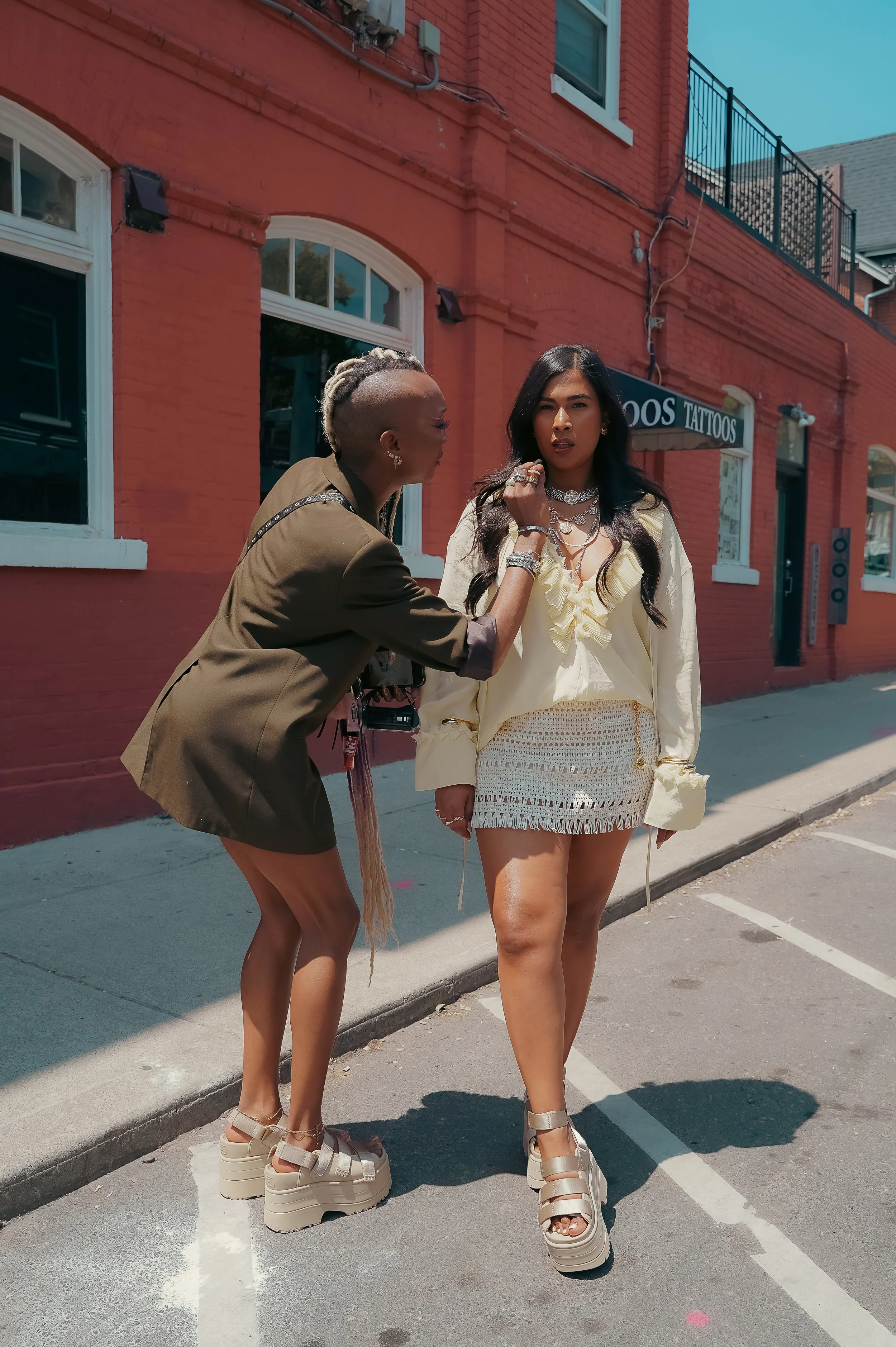 Two women standing on a city street during daytime, one appears to be touching the other's face, both wearing fashionable outfits and chunky platform sandals, with a red building and tattoo shop sign in the background.