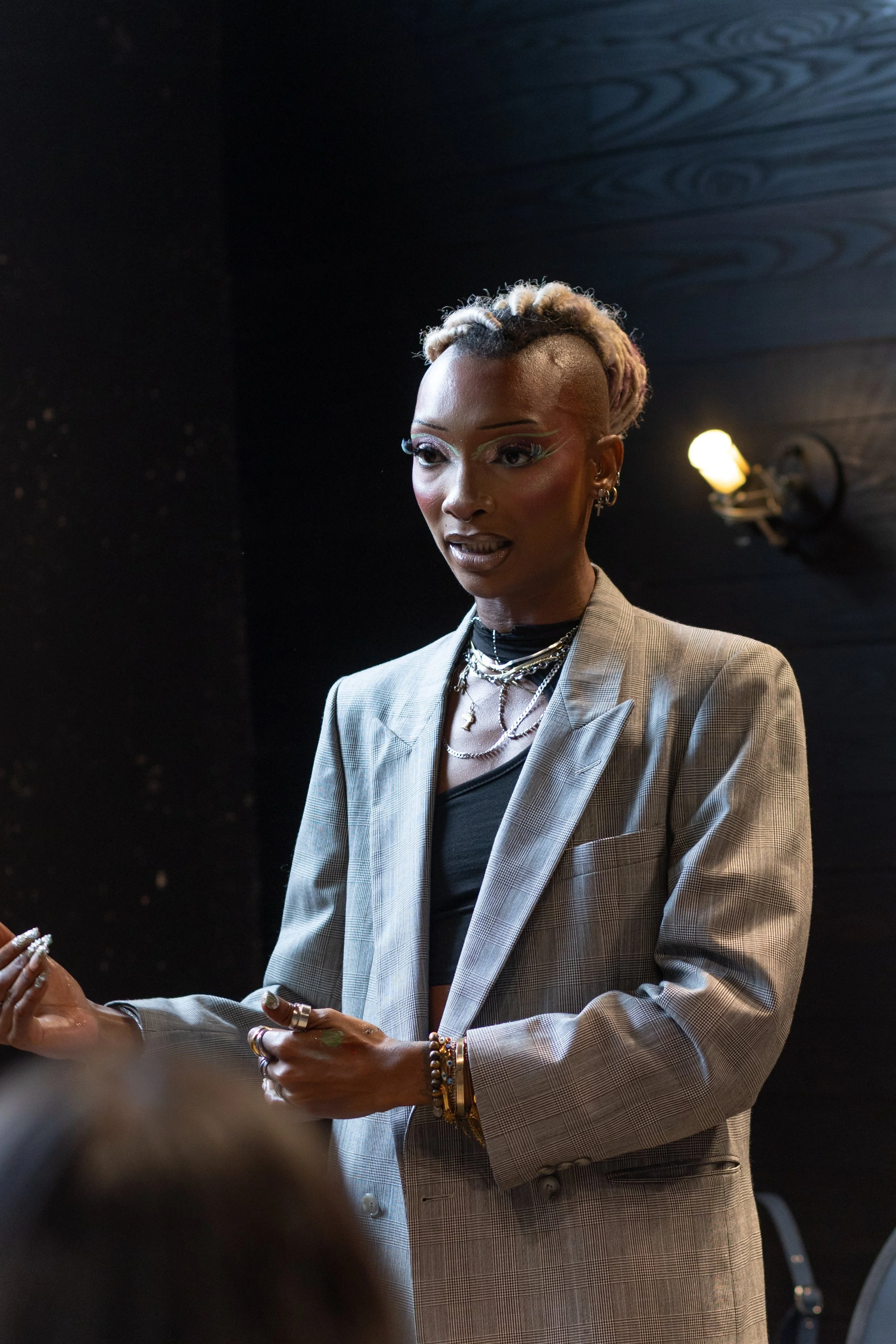 A woman with styled hair, wearing a plaid blazer, layered necklaces, and bracelets, standing in front of a dark background with a wall-mounted light.