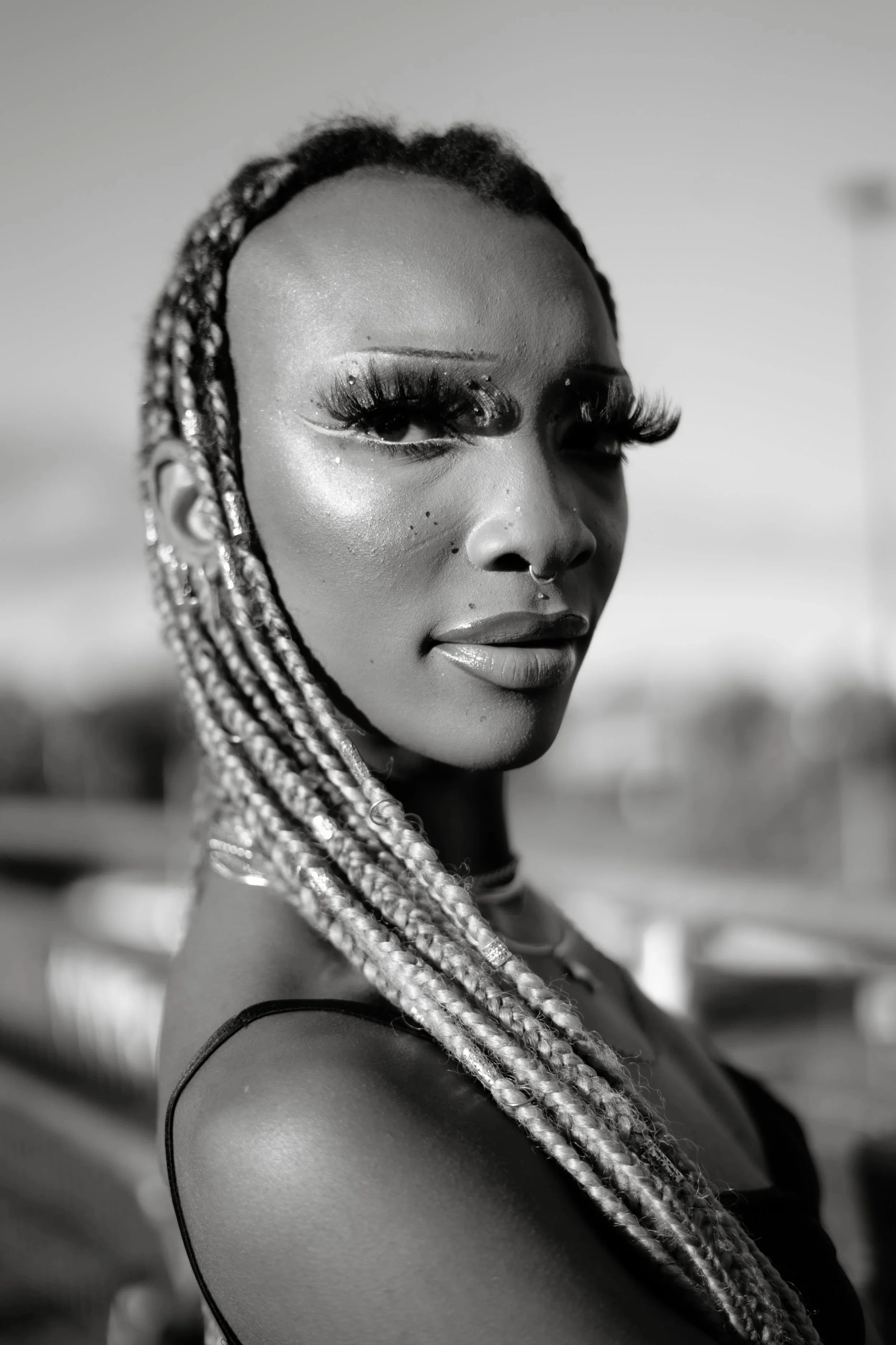 Black and white portrait of a woman with braided hair, glamorous makeup with long lashes, and a septum piercing, looking at the camera.