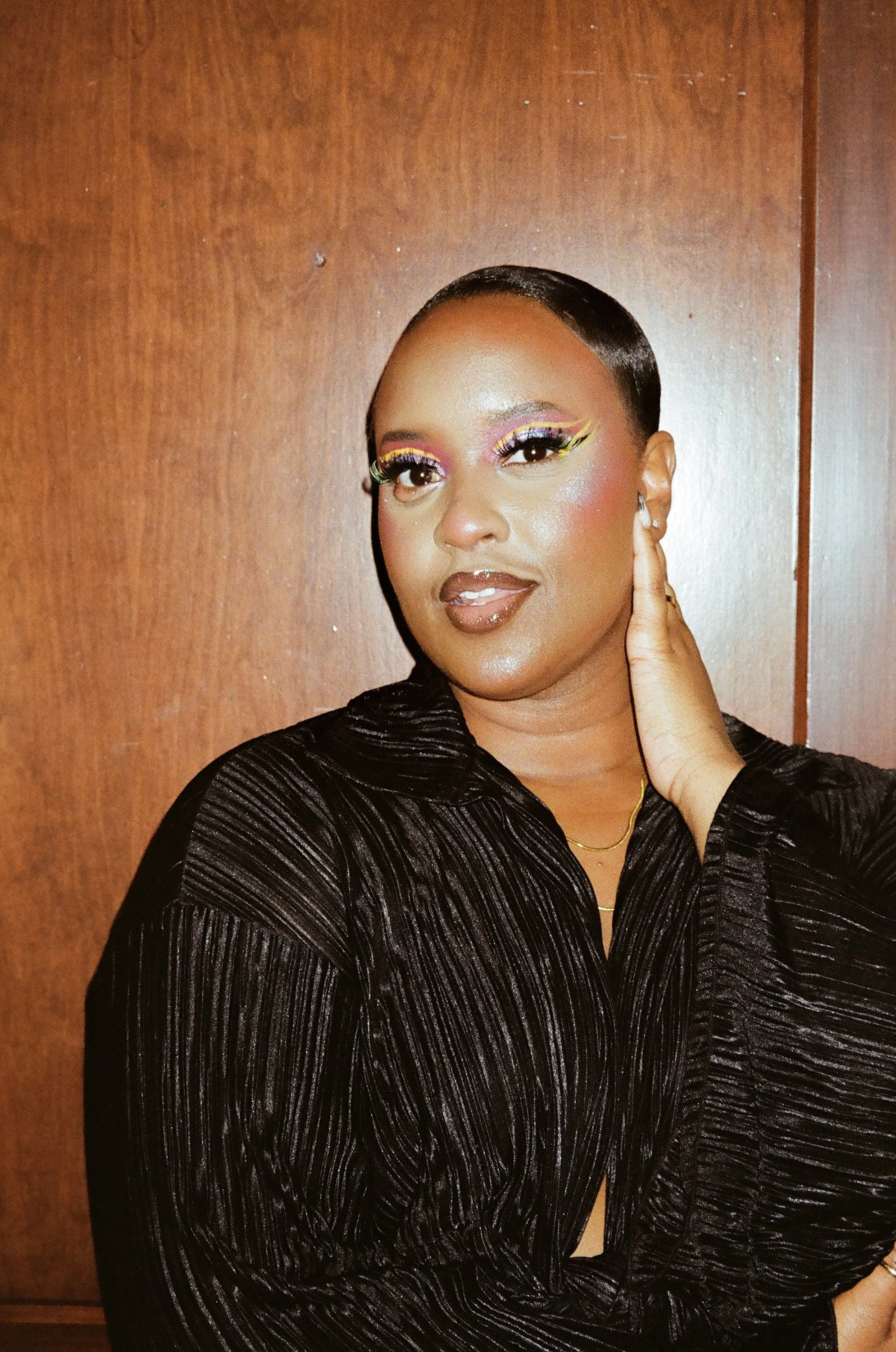 A woman with short hair and colorful eye makeup, wearing a black velvet dress, standing against a wooden wall.