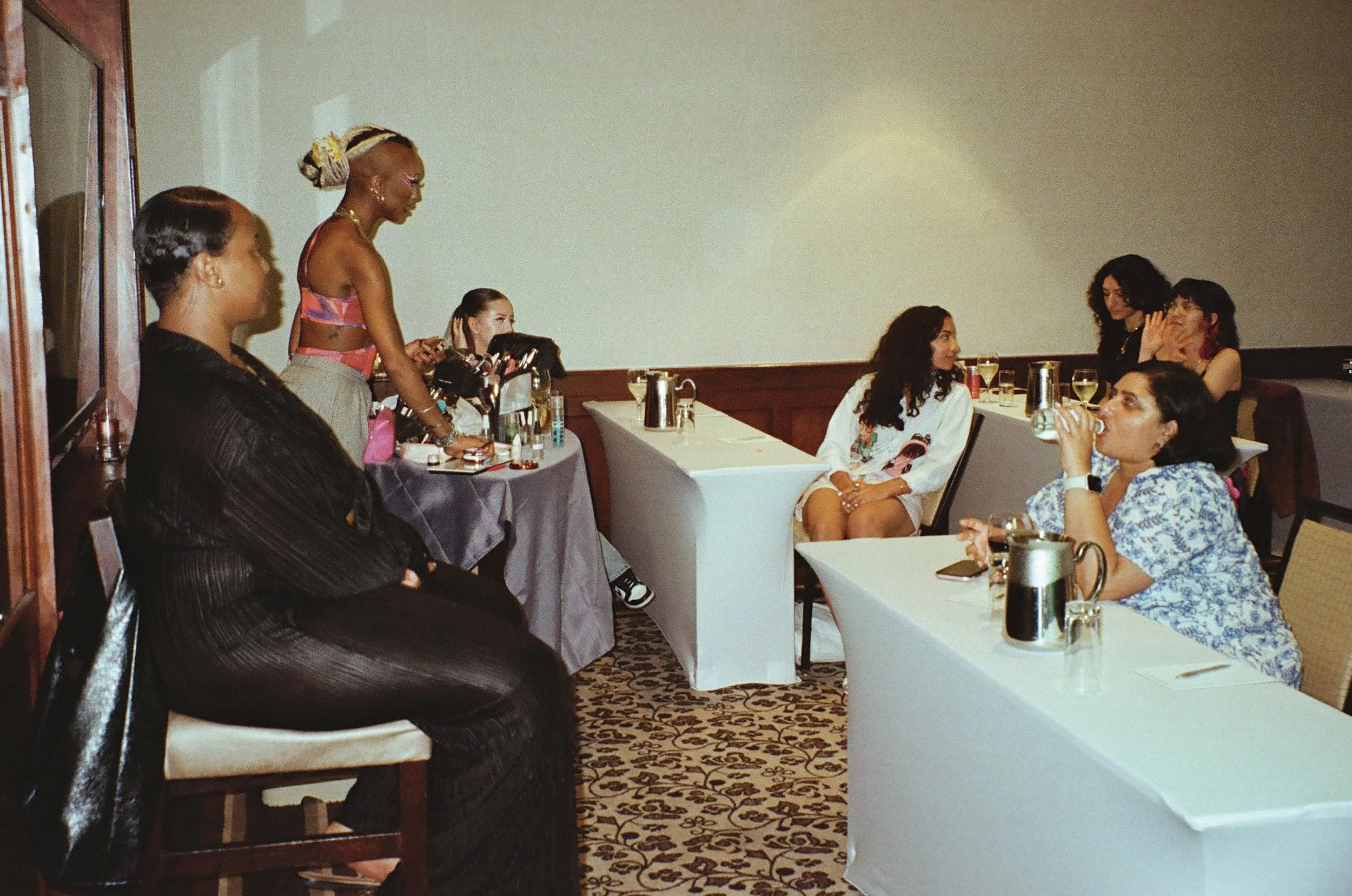 A group of women attending a makeup demonstration or tutorial in a conference room, some are seated at tables with drinks, while one woman stands at a table with makeup products.
