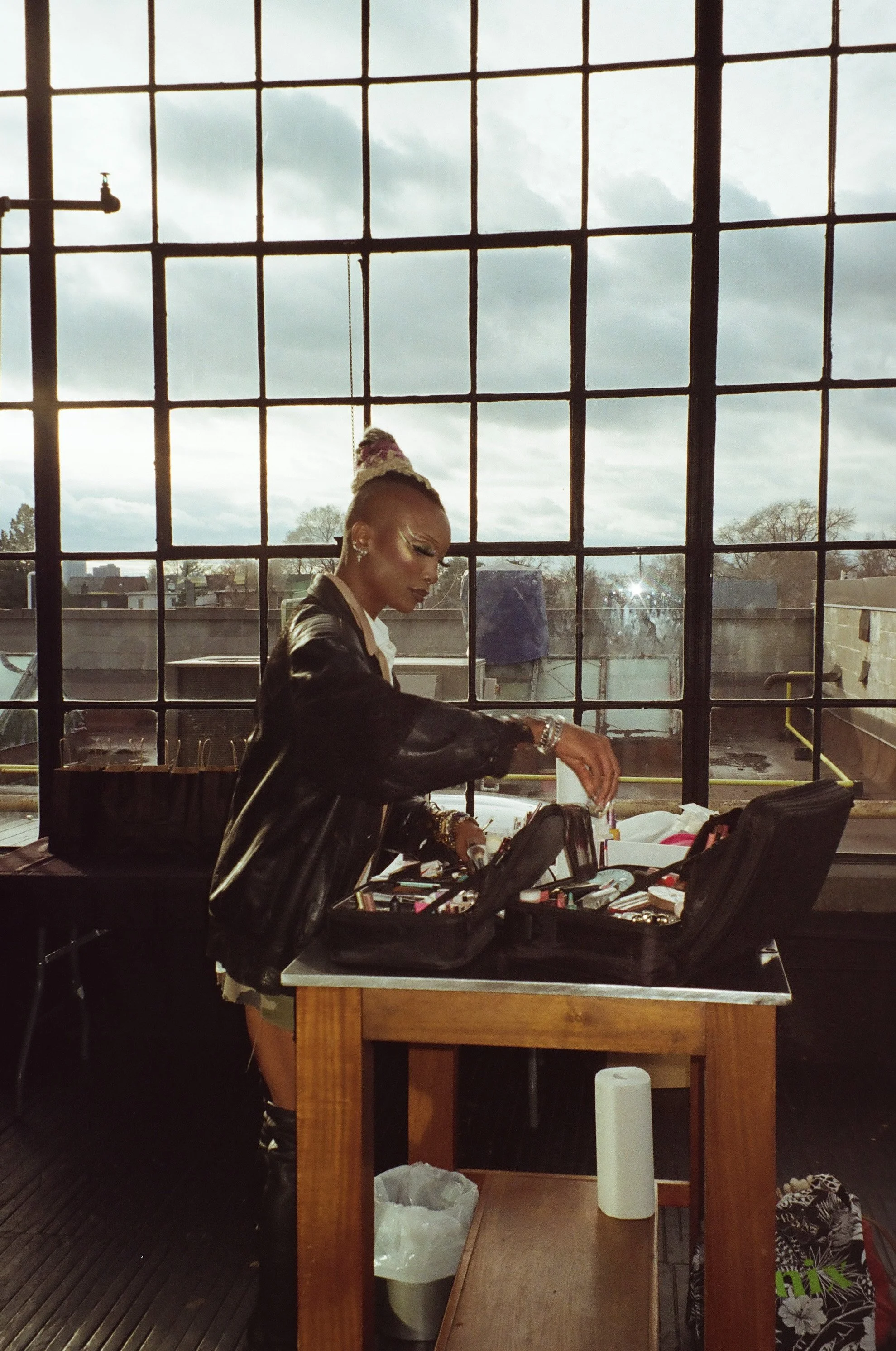 A woman with colorful braided hair and makeup is organizing makeup and accessories at a table near large industrial-style windows with a cloudy sky outside.
