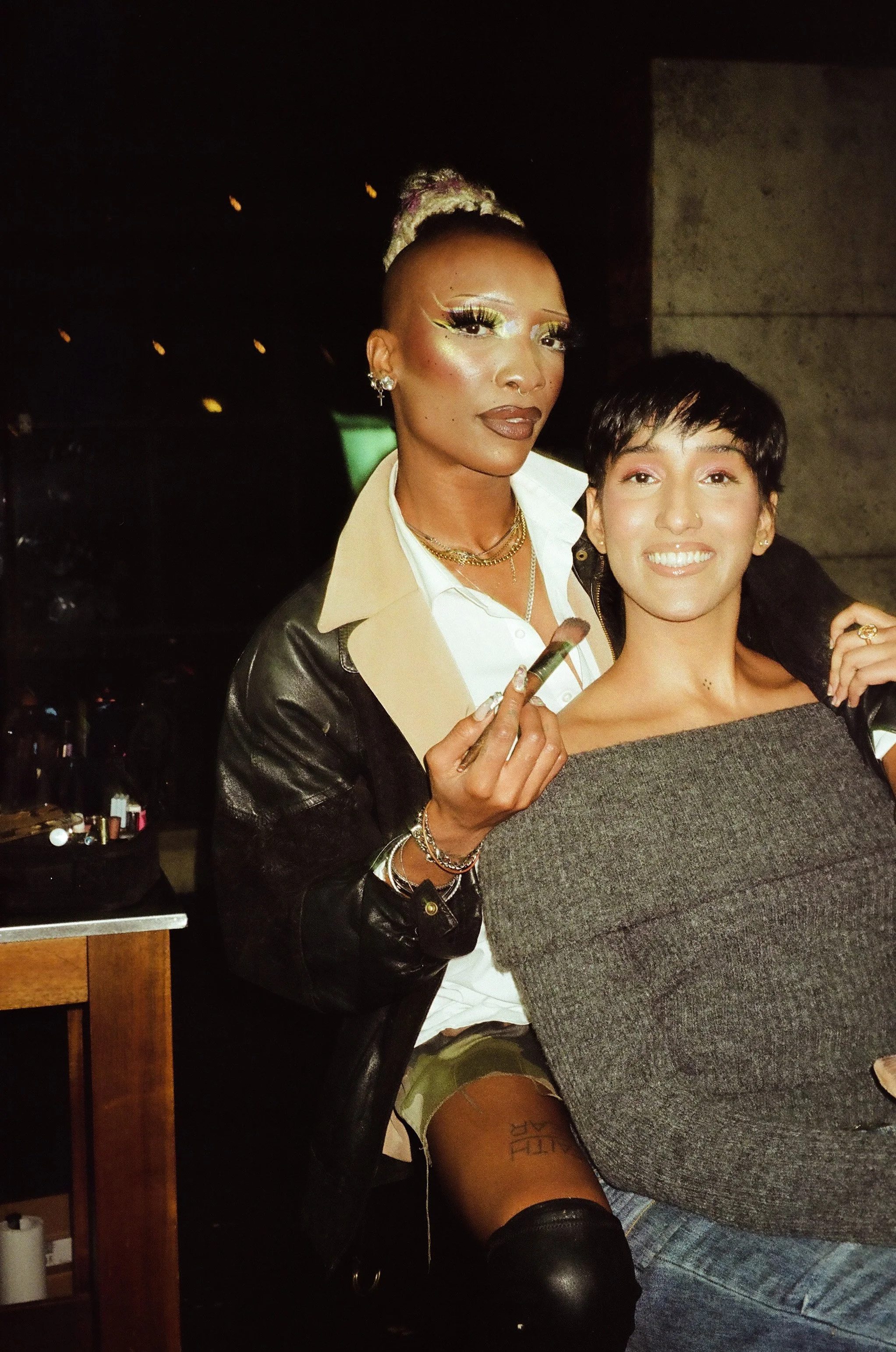 Two women, one with bold makeup and jewelry, holding a makeup brush, sitting closely together and smiling in a dimly lit setting.